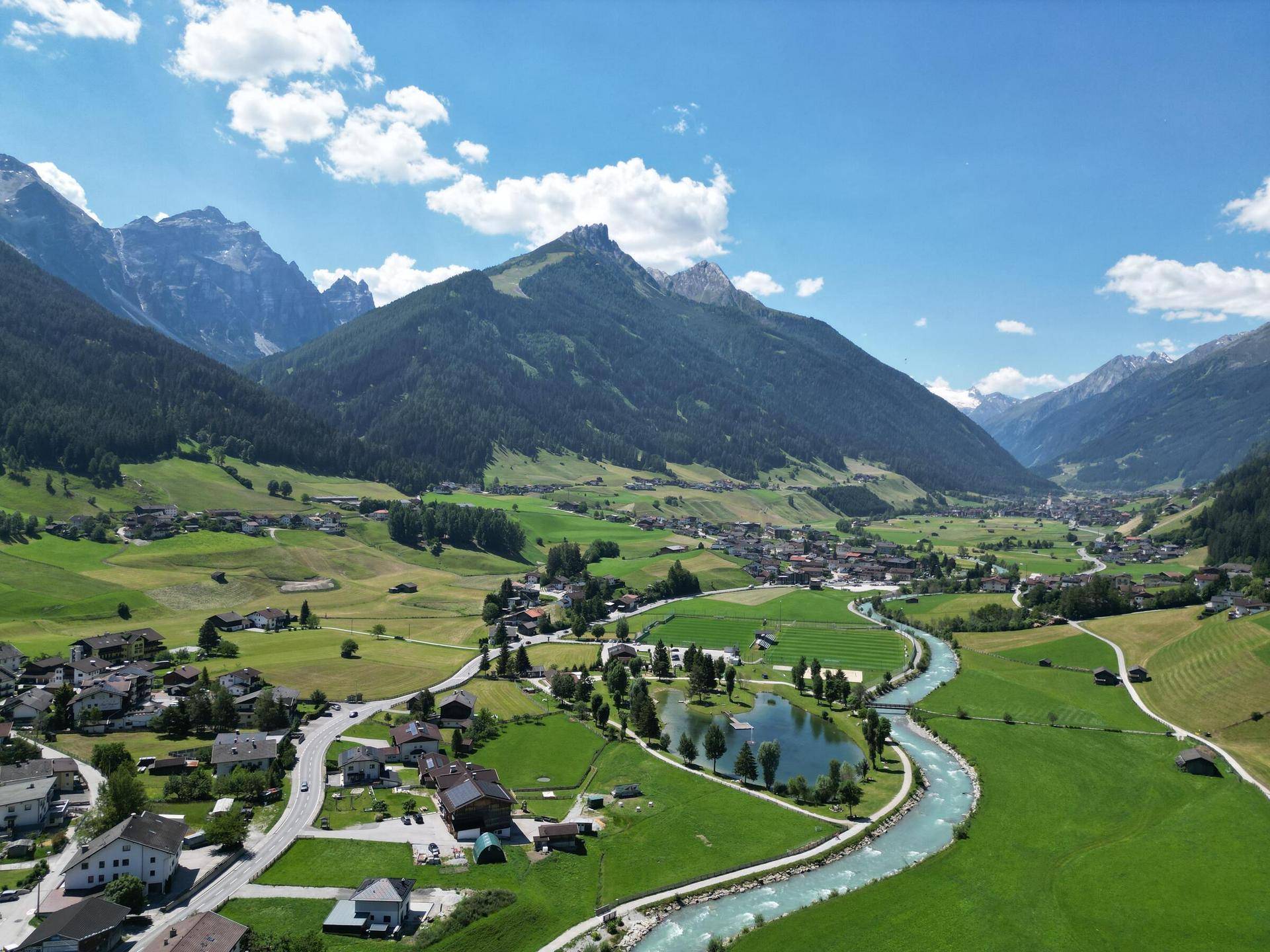 Atemberaubende Luftaufnahme eines grünen Tals mit einem Fluss, See und kleinen Siedlungen, umgeben von hohen Bergen.