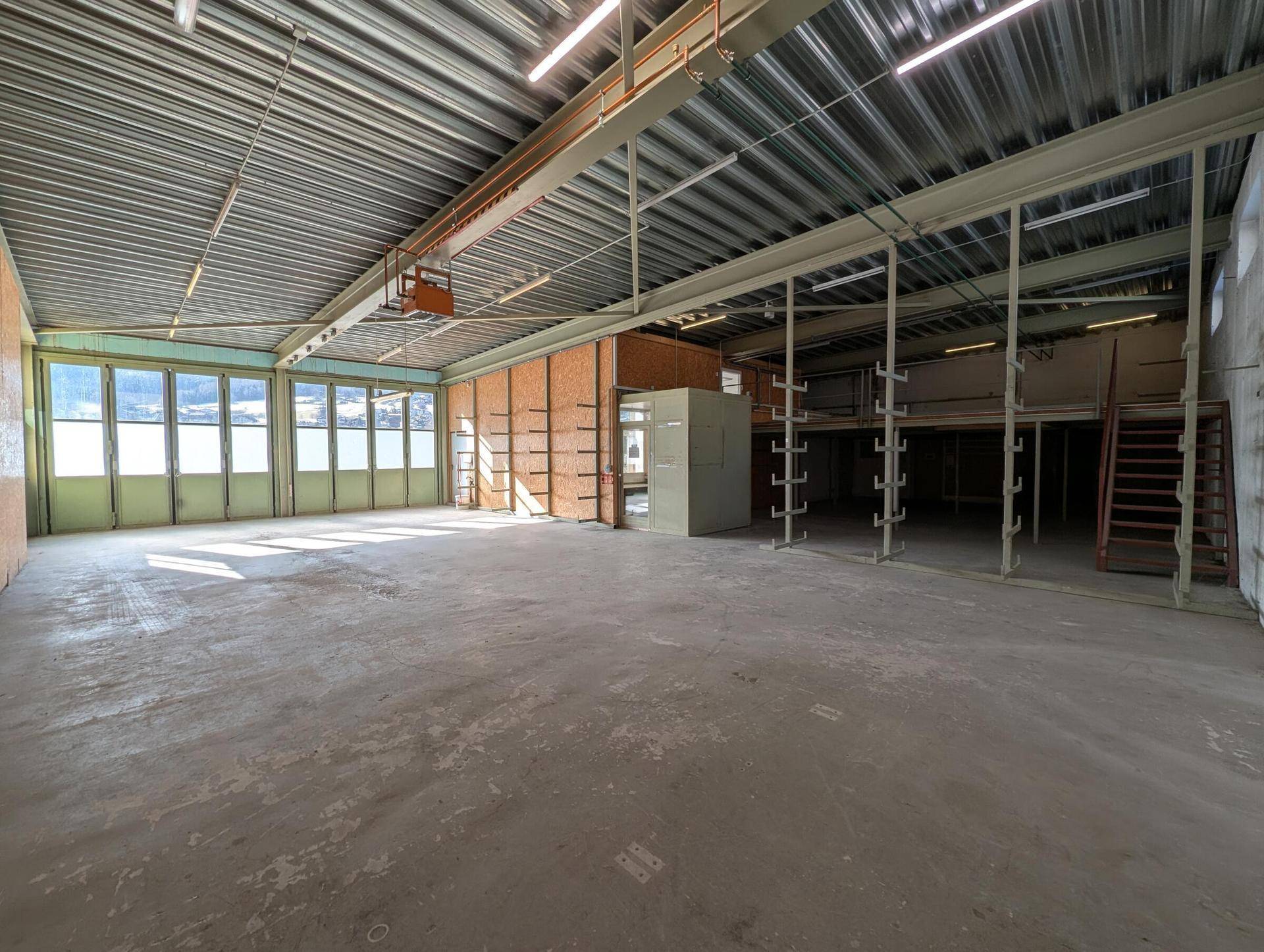 Geräumige Lagerhalle mit hoher Decke, Betonboden und großen Fenstern, die viel Tageslicht hereinlassen.