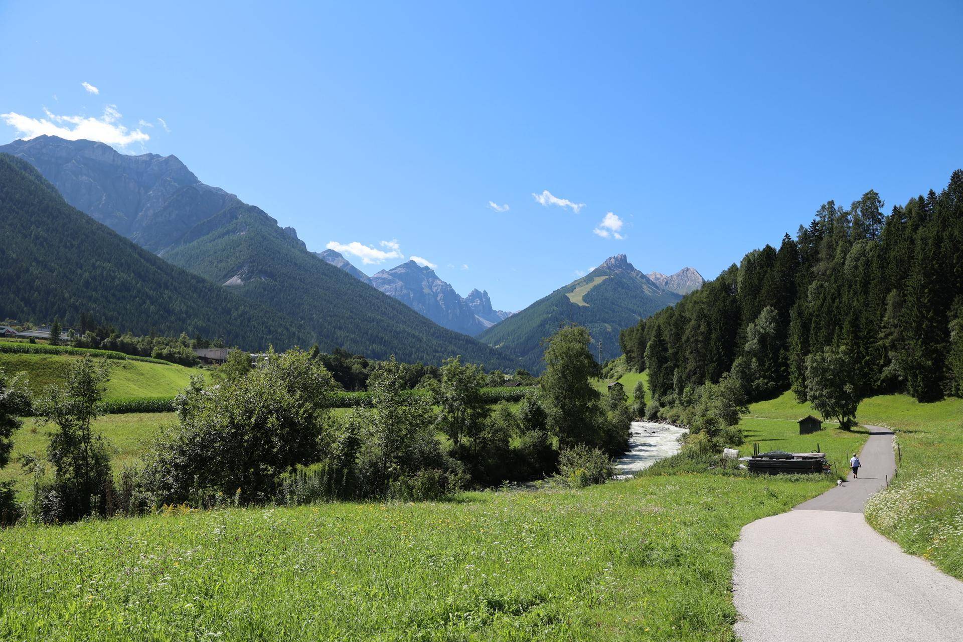 Malerische Landschaft mit grünen Wiesen, einem Fluss und majestätischen Bergen unter blauem Himmel.