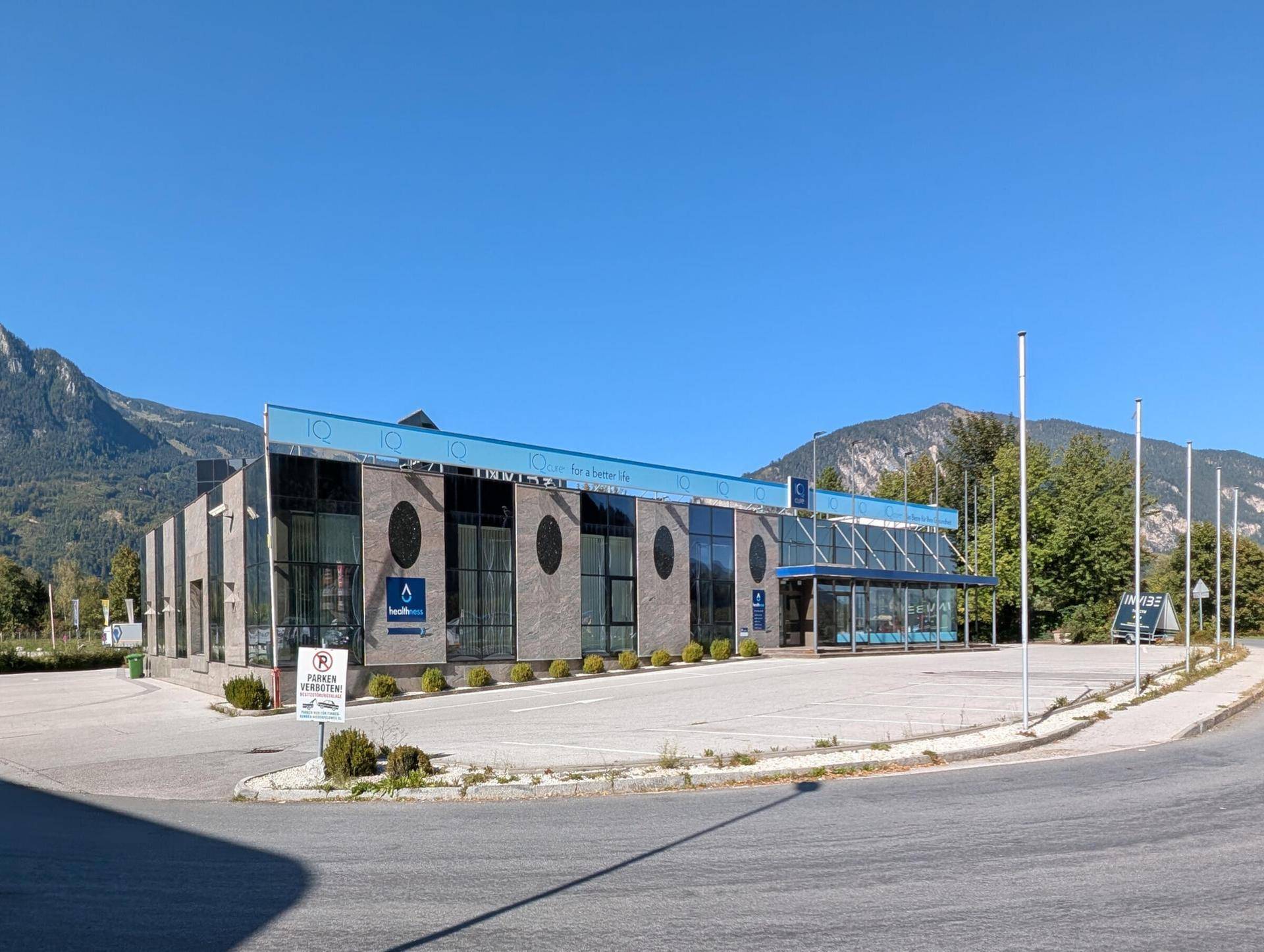Modernes Geschäftsgebäude mit Glasfassade, Parkplätzen und Bergblick unter blauem Himmel.