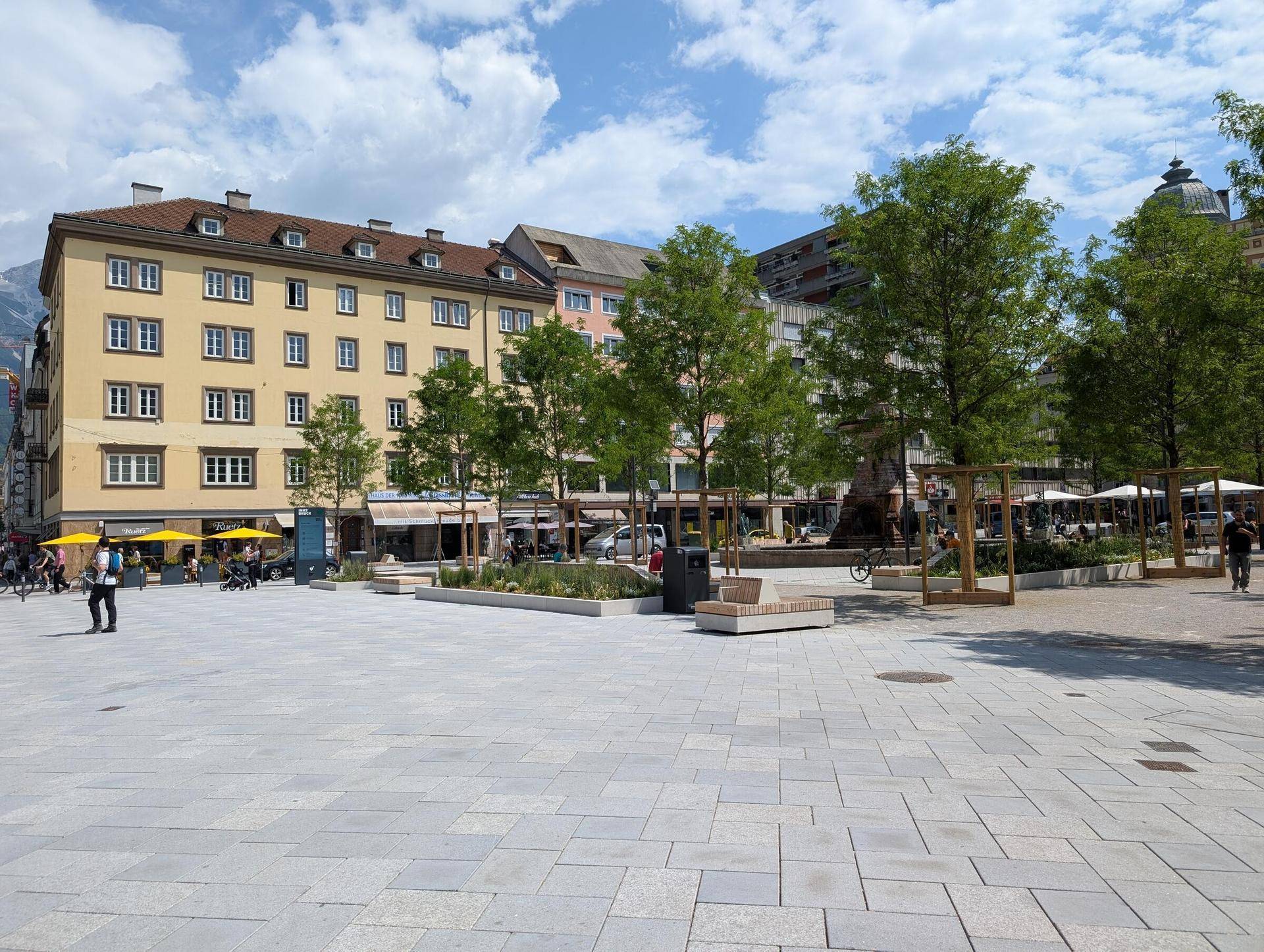 Weitläufiger Stadtplatz mit gepflastertem Boden, umgeben von Gebäuden, Bäumen und Außengastronomie.