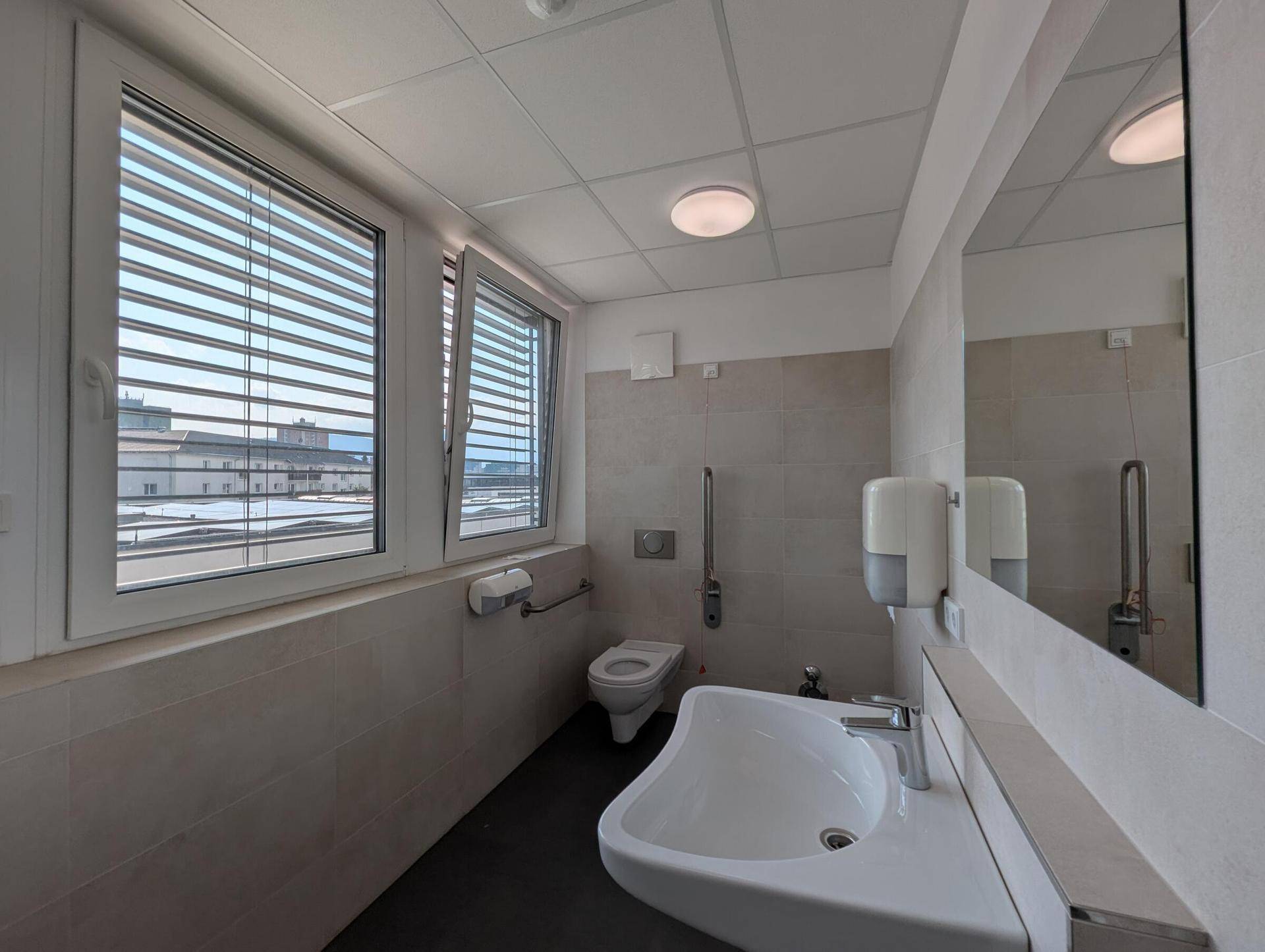 Modernes Badezimmer mit Fliesenboden, Fenster und barrierefreien Haltegriffen, ausgestattet mit einem Waschbecken.