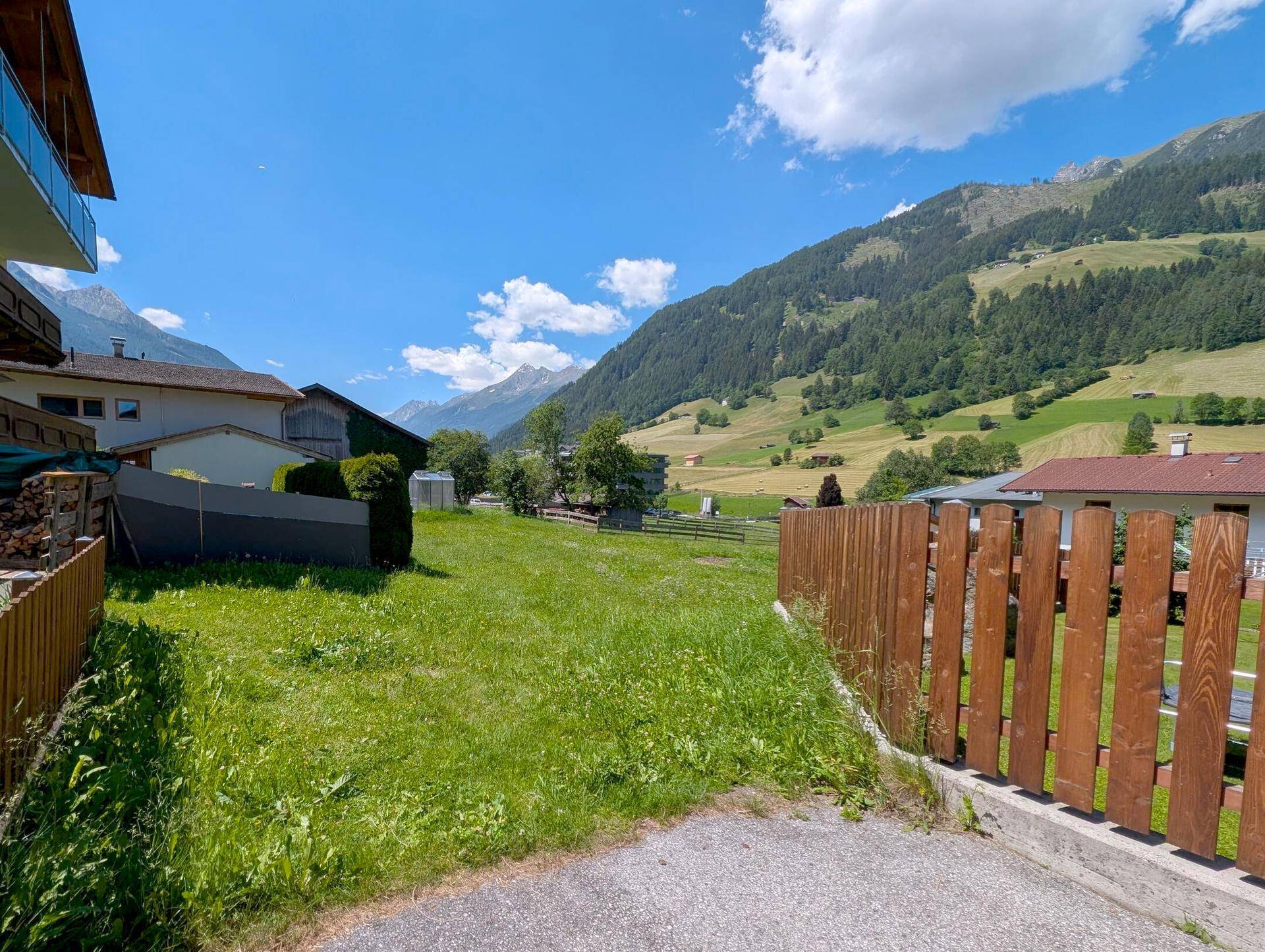 Weitläufiger Rasenbereich mit Holzzaun und Häusern, umgeben von einer grünen Berglandschaft unter strahlend blauem Himmel.