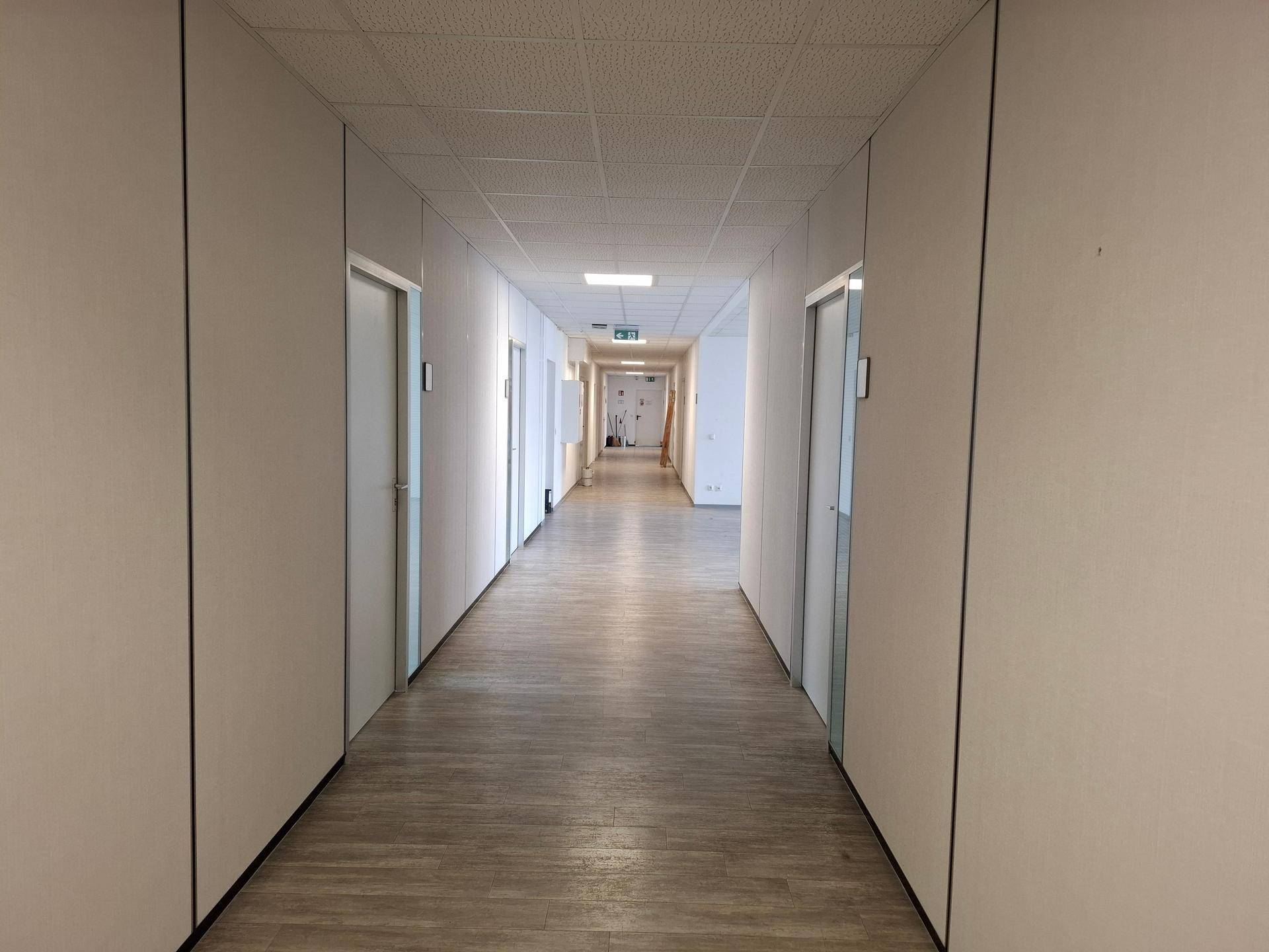 Langer Bürogang mit hellem Laminatboden und weißen Trennwänden, der zu weiteren Räumen führt.