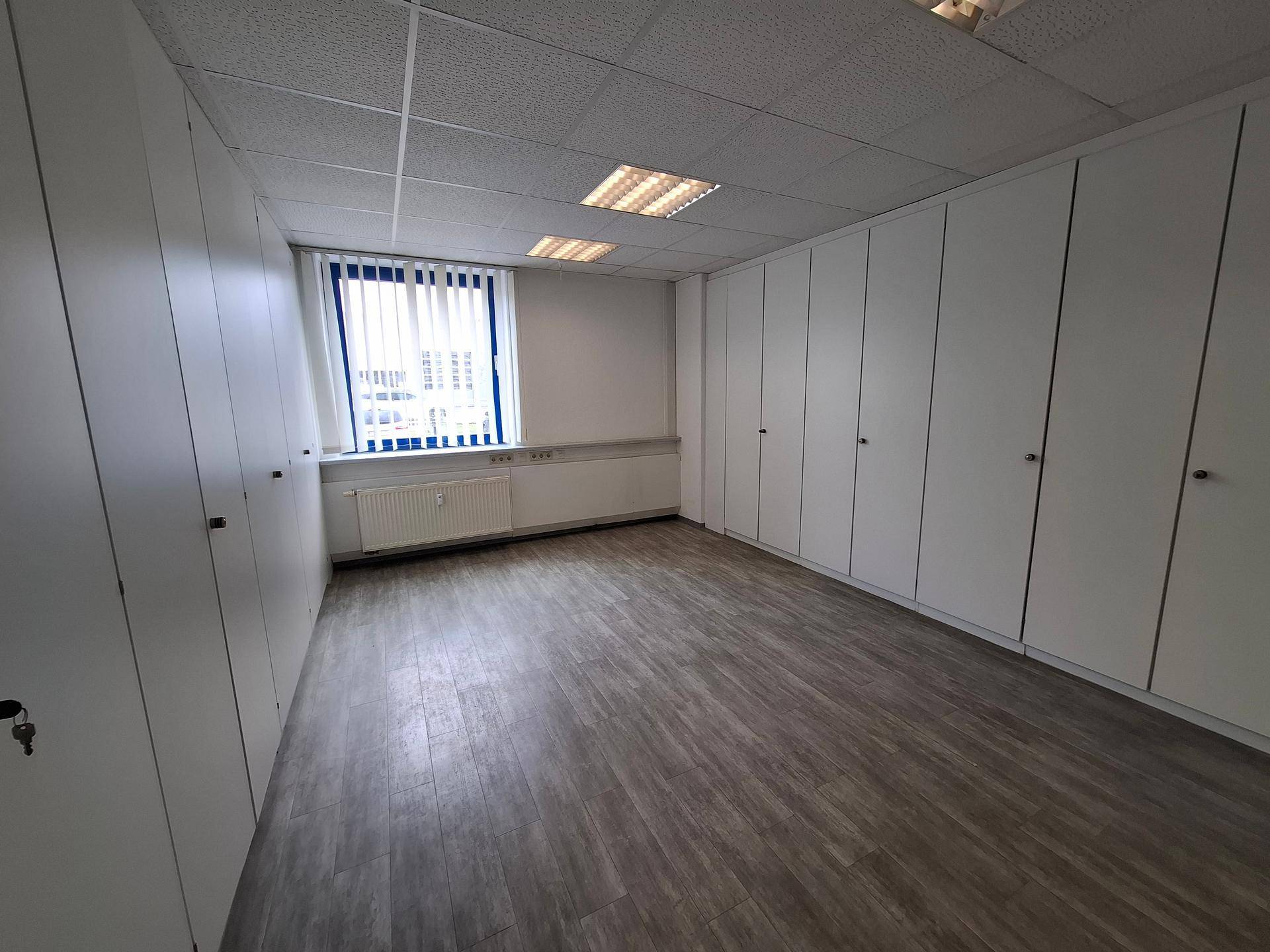 Büroraum mit Laminatboden, weißen Wänden, Fenster mit Jalousien und integrierten Schränken.