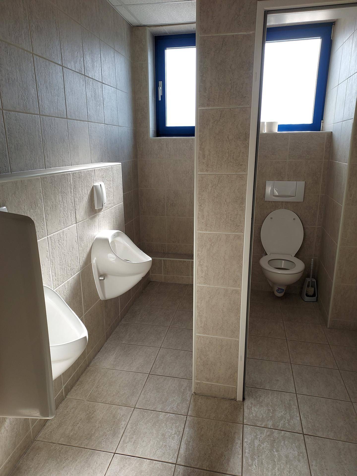 Herren-WC mit zwei Urinalen, einer Toilettenkabine und einem Fenster für Tageslicht.