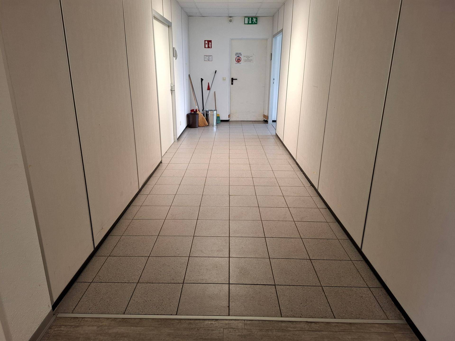Ende eines Bürogangs mit Fliesenboden und weißen Wänden, Abstellbereich für Reinigungsgeräte.