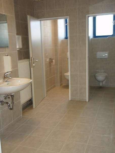Sanitärraum mit Waschbecken, Heizkörper und zwei Toilettenkabinen, hell gefliest.