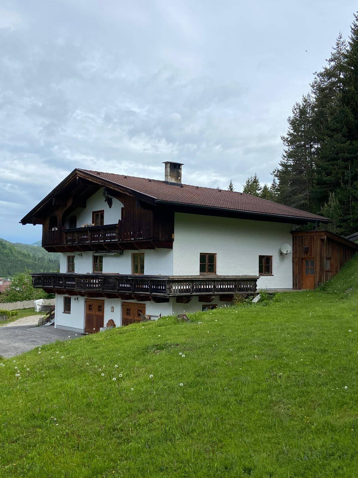 Traditionelles Alpenhaus mit weißer Fassade, Holzbalkonen und Satteldach, eingebettet in eine grüne Hügellandschaft.