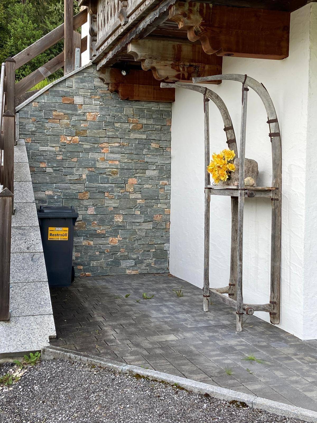 Rustikale Außendekoration mit einem alten Holzschlitten und Blumen vor einer Steinmauer und gepflastertem Bereich.