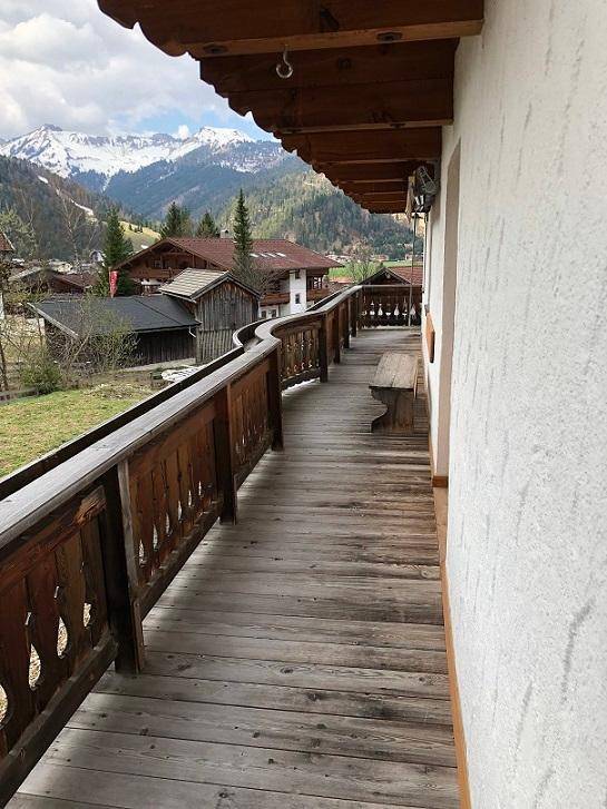 Langer Holzbalkon mit traditionellem Geländer und malerischem Blick auf die umliegenden Berge.
