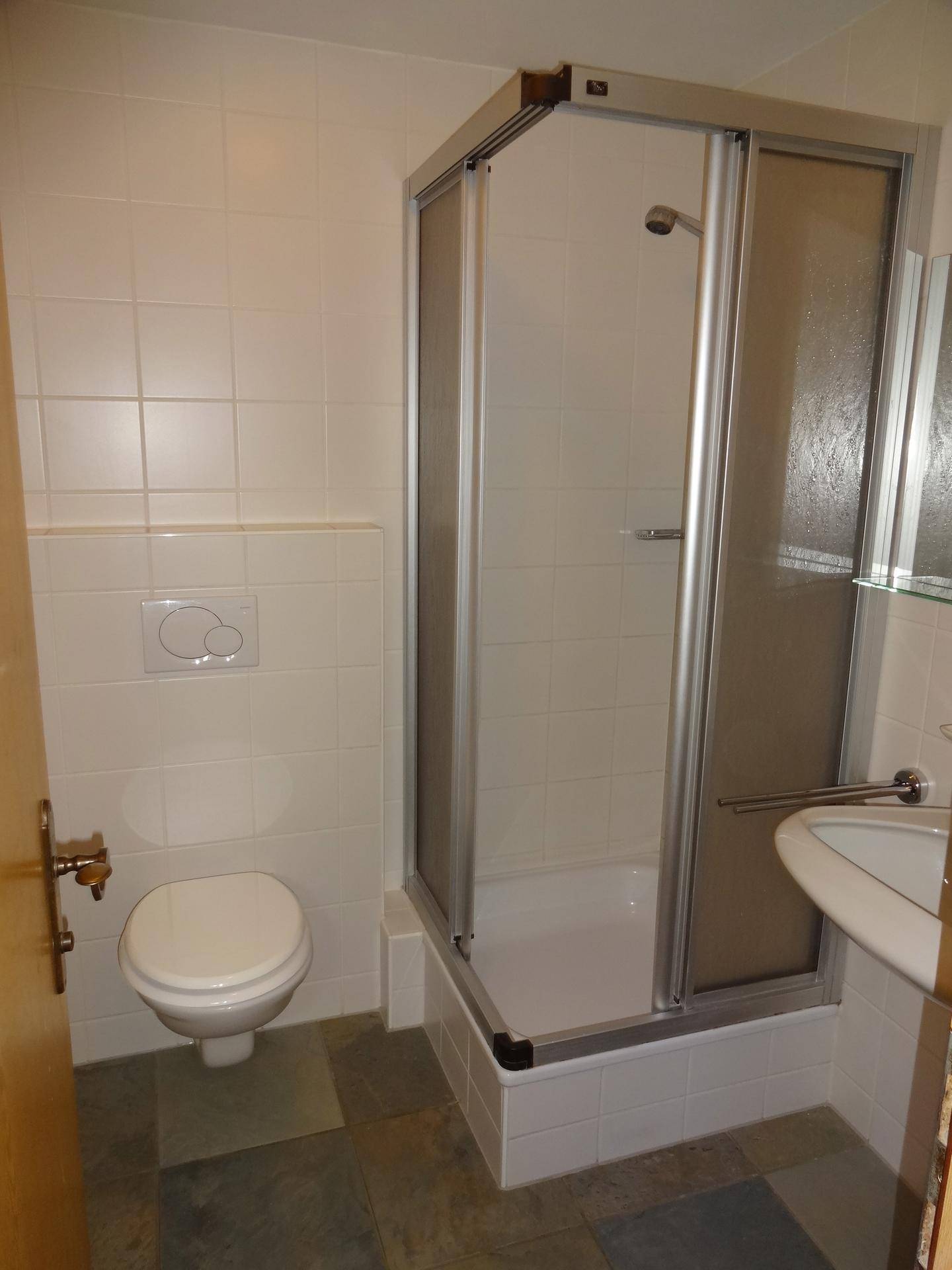Badezimmer mit Dusche, WC und Waschbecken, gefliest in Weiß und Grau.