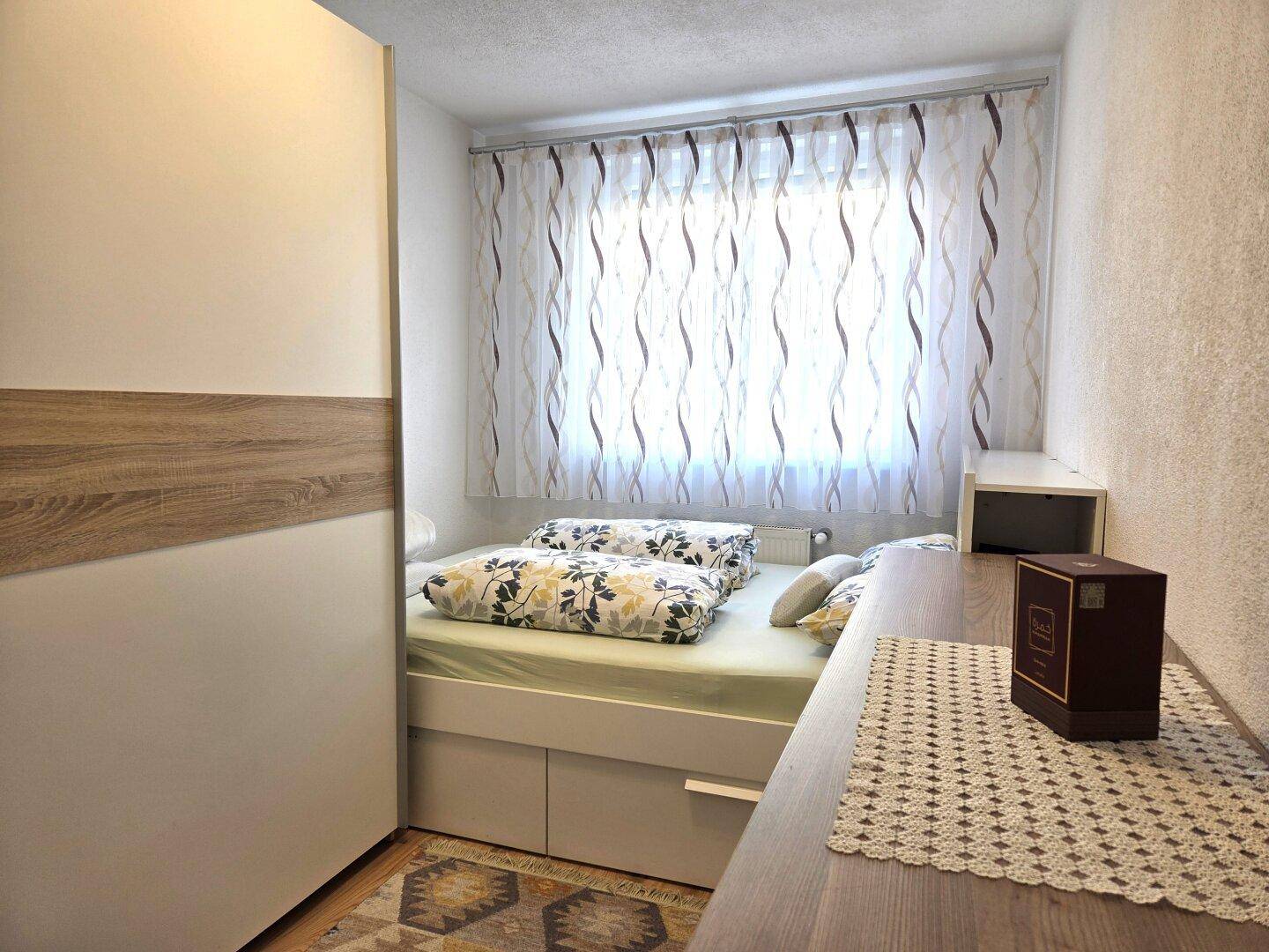 Helles Schlafzimmer mit Bett, Kleiderschrank und Fenster mit Vorhängen.