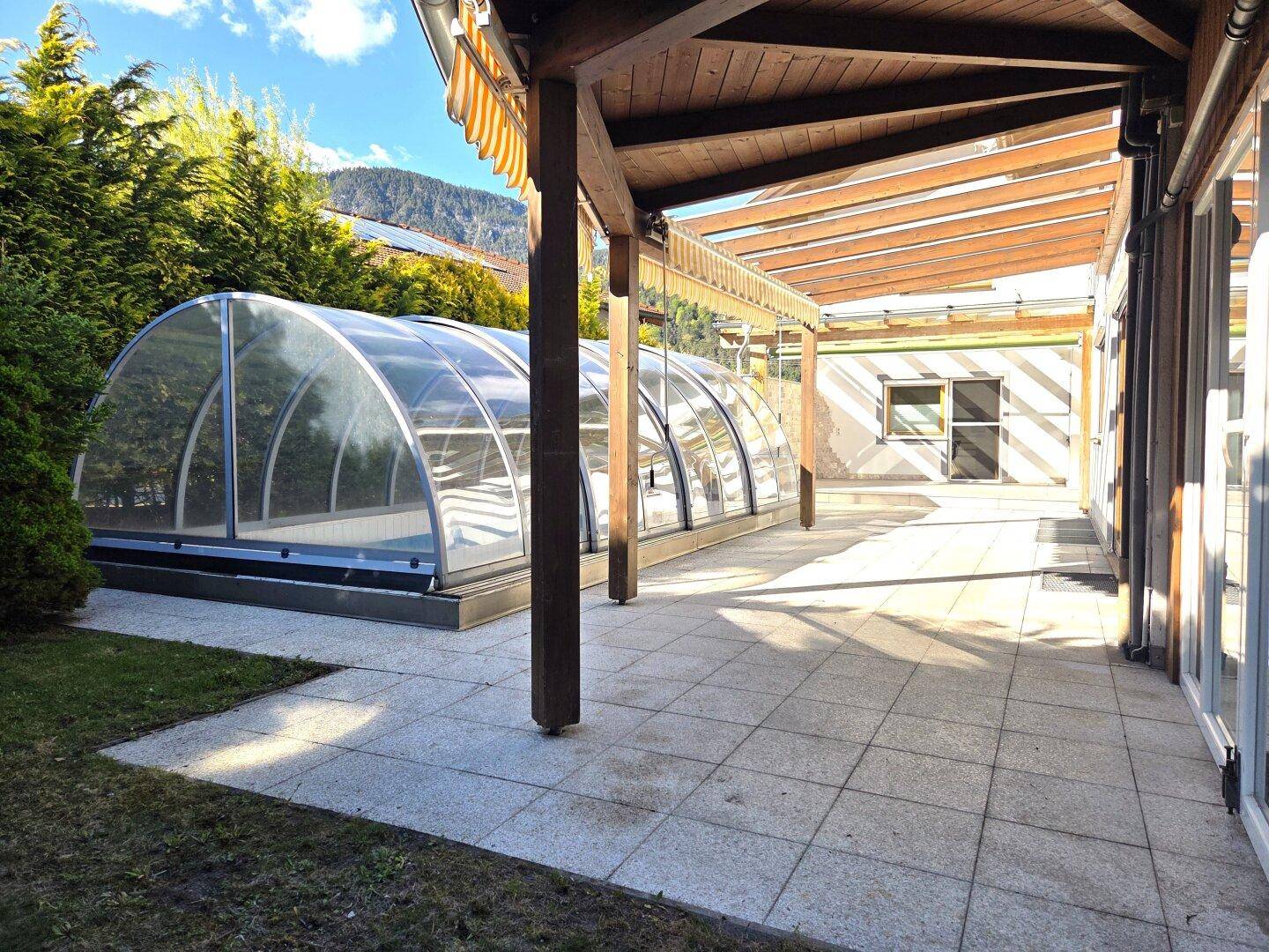 Sonniger Terrassenbereich mit überdachtem Pool und Holzbalkenkonstruktion, umgeben von Grün.