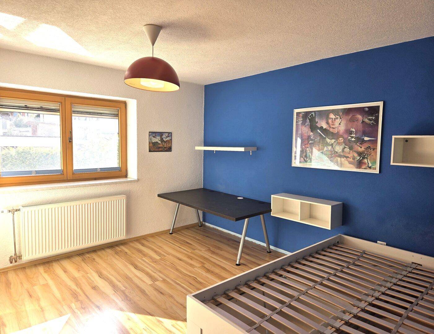 Kinderzimmer mit blau gestrichener Wand, Holzboden und einem Bettgestell ohne Matratze.