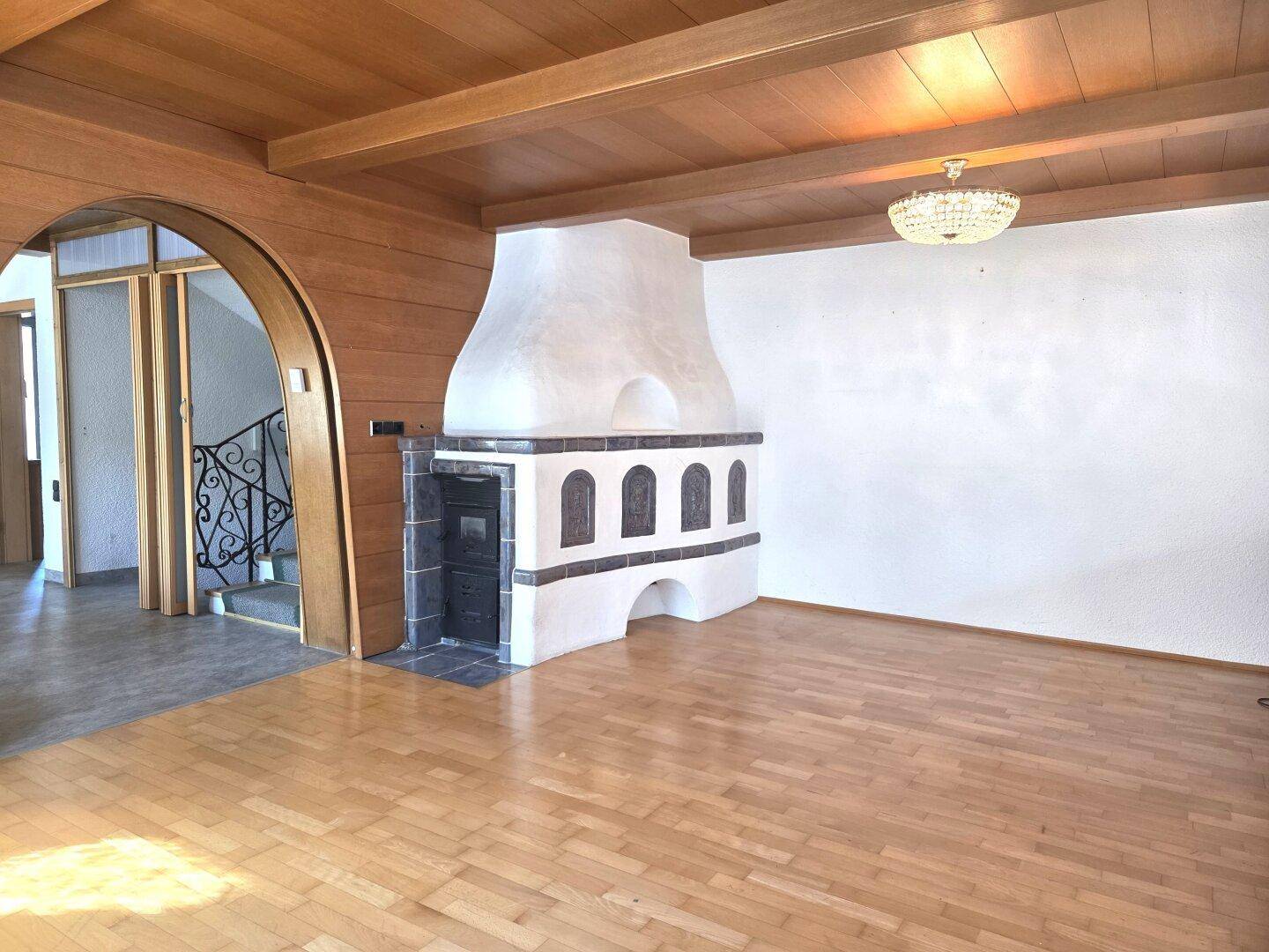 Geräumiges Wohnzimmer mit Holzbalkendecke, Parkettboden und einem traditionellen Kachelofen.