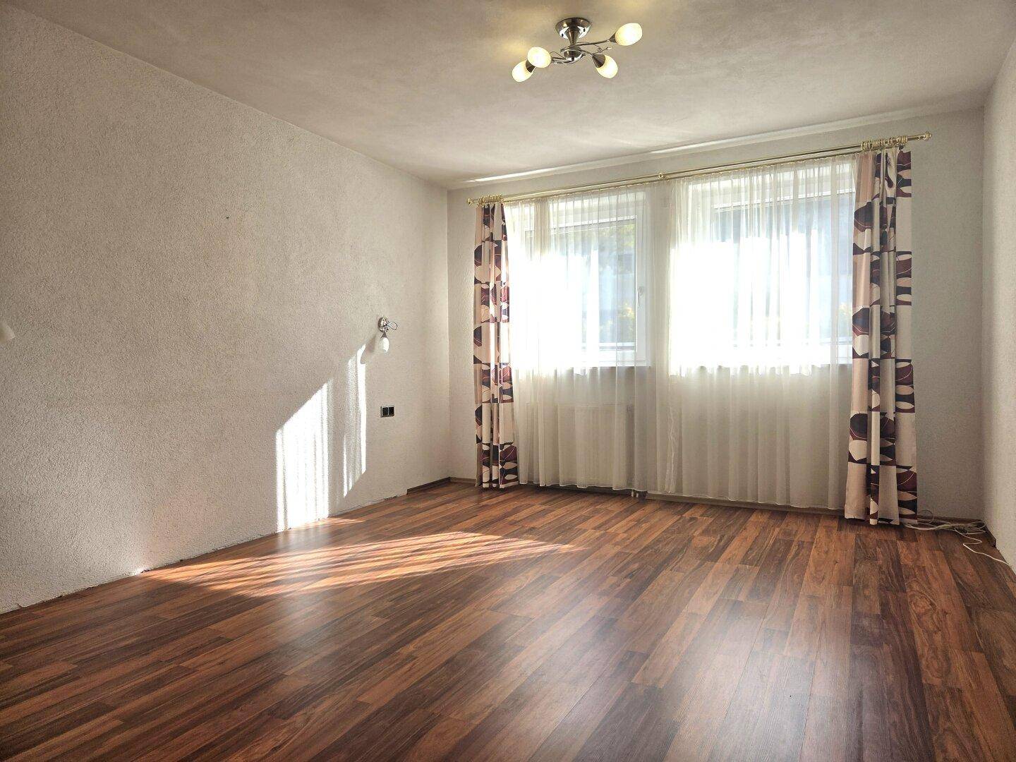 Helles Wohnzimmer mit dunklem Laminatboden, großen Fenstern und Vorhängen.