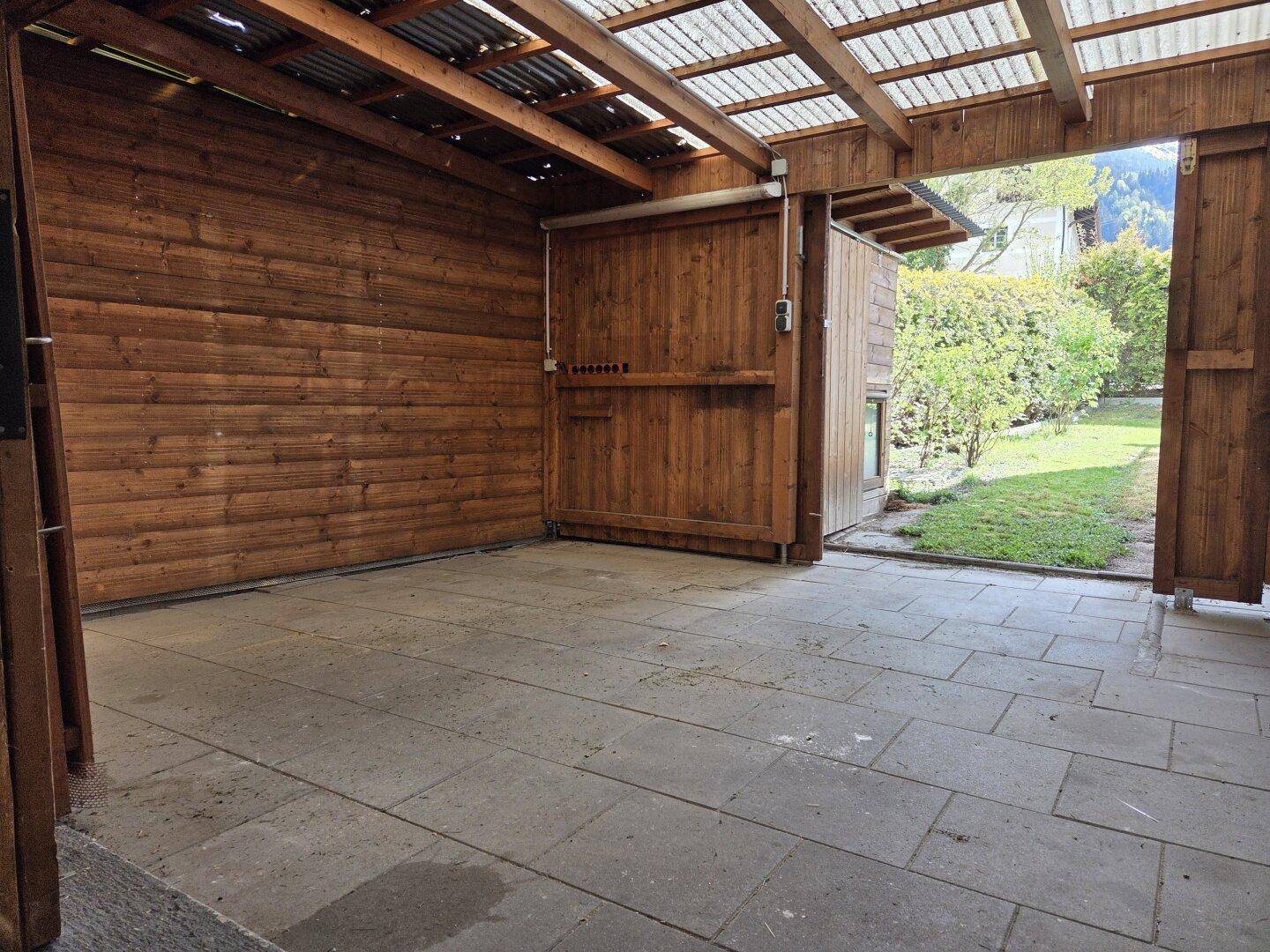 Überdachter Stellplatz oder Carport mit Holzwänden und gepflastertem Boden.