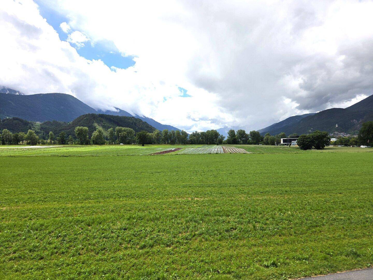 Weitläufige grüne Felder und Wiesen mit einer beeindruckenden Bergkulisse unter bewölktem Himmel.