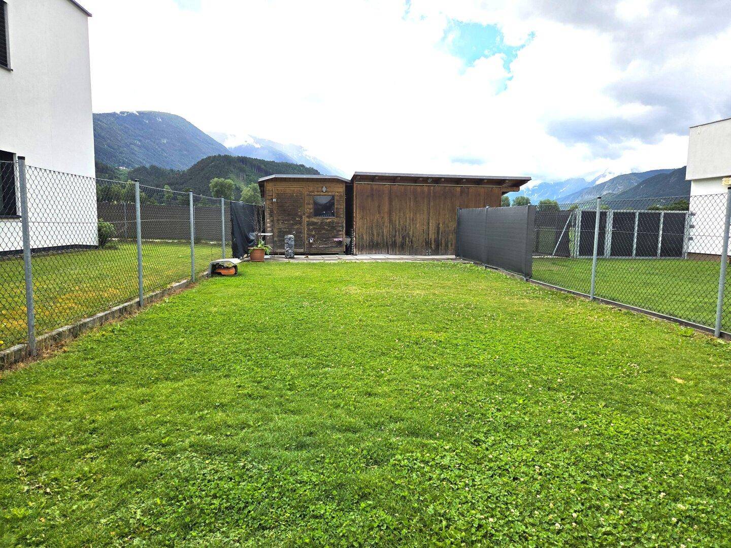 Eingezäunter Gartenbereich mit Rasenfläche, zwei Holzschuppen und Blick auf die Berge im Hintergrund.