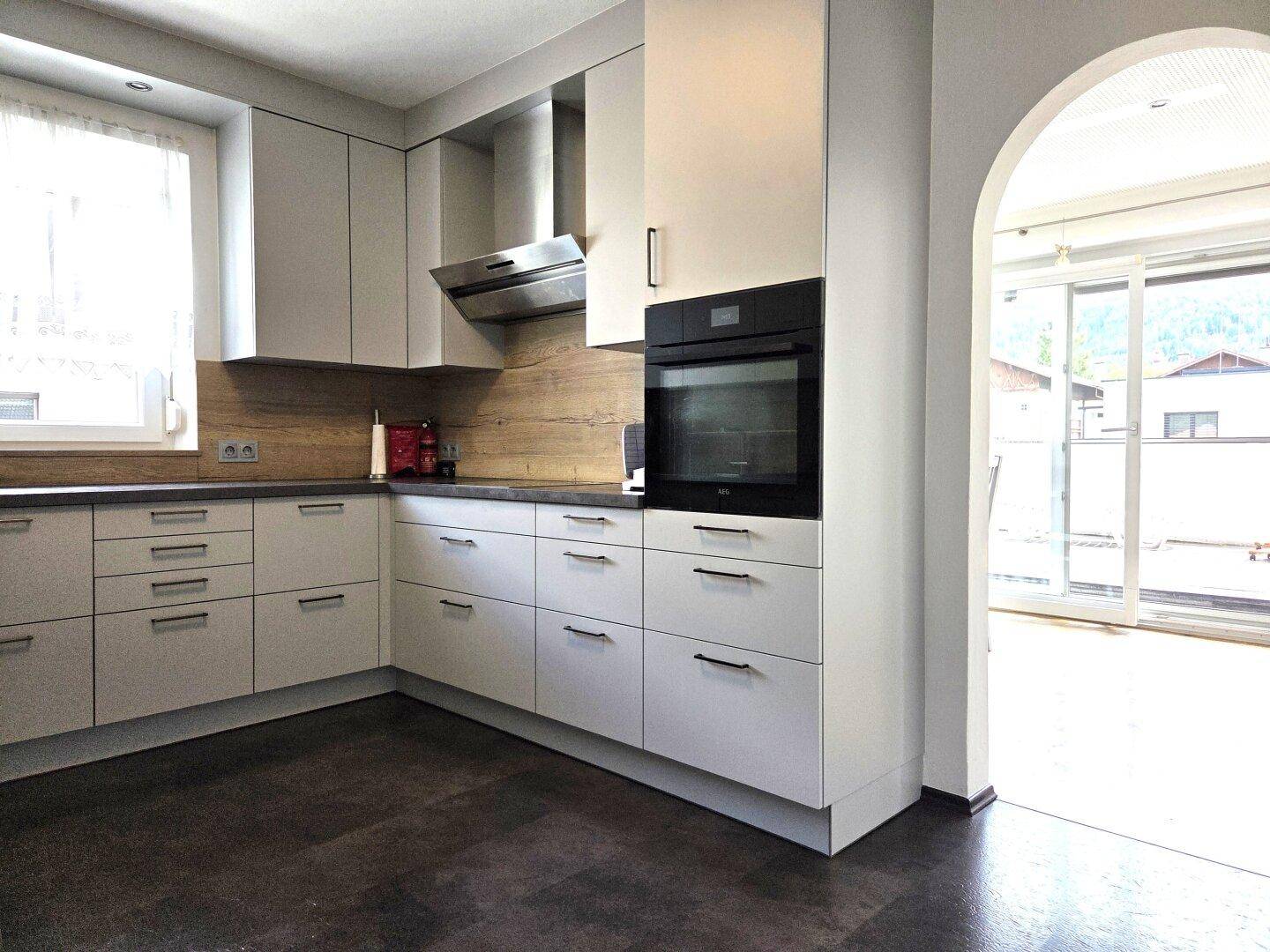 Moderne Einbauküche mit weißen Fronten, Holzdekor und integriertem Backofen, offen zu einem weiteren Raum.