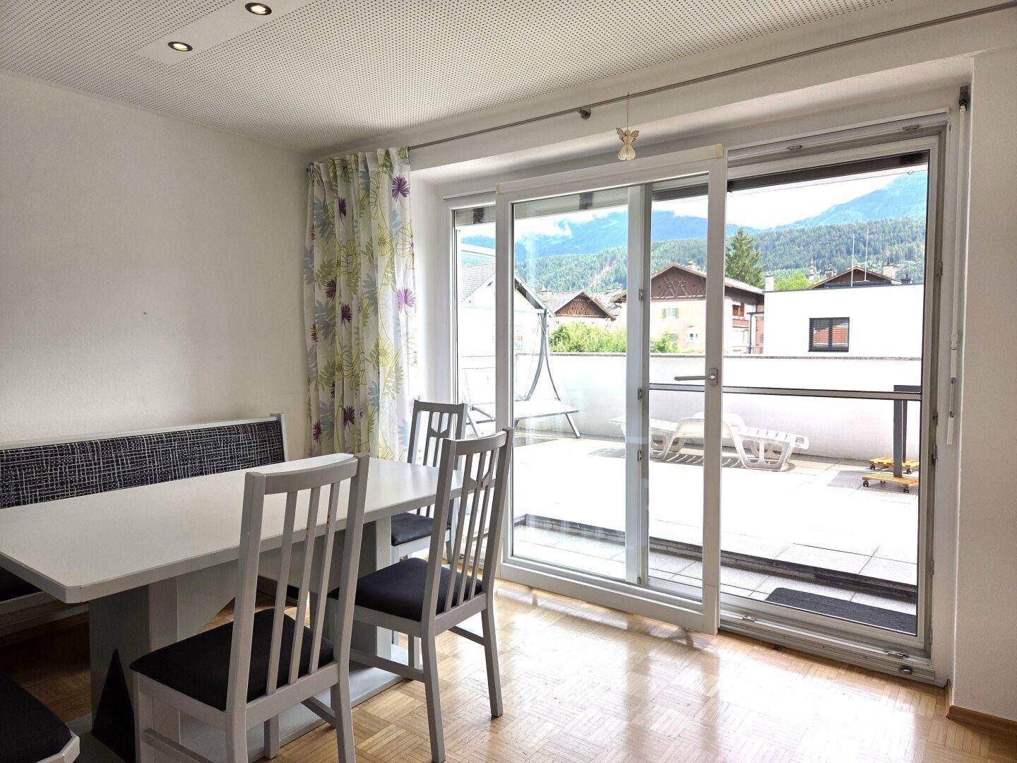 Essbereich mit weißem Esstisch, Sitzbank und großzügigem Zugang zu einer sonnigen Terrasse mit Bergblick.