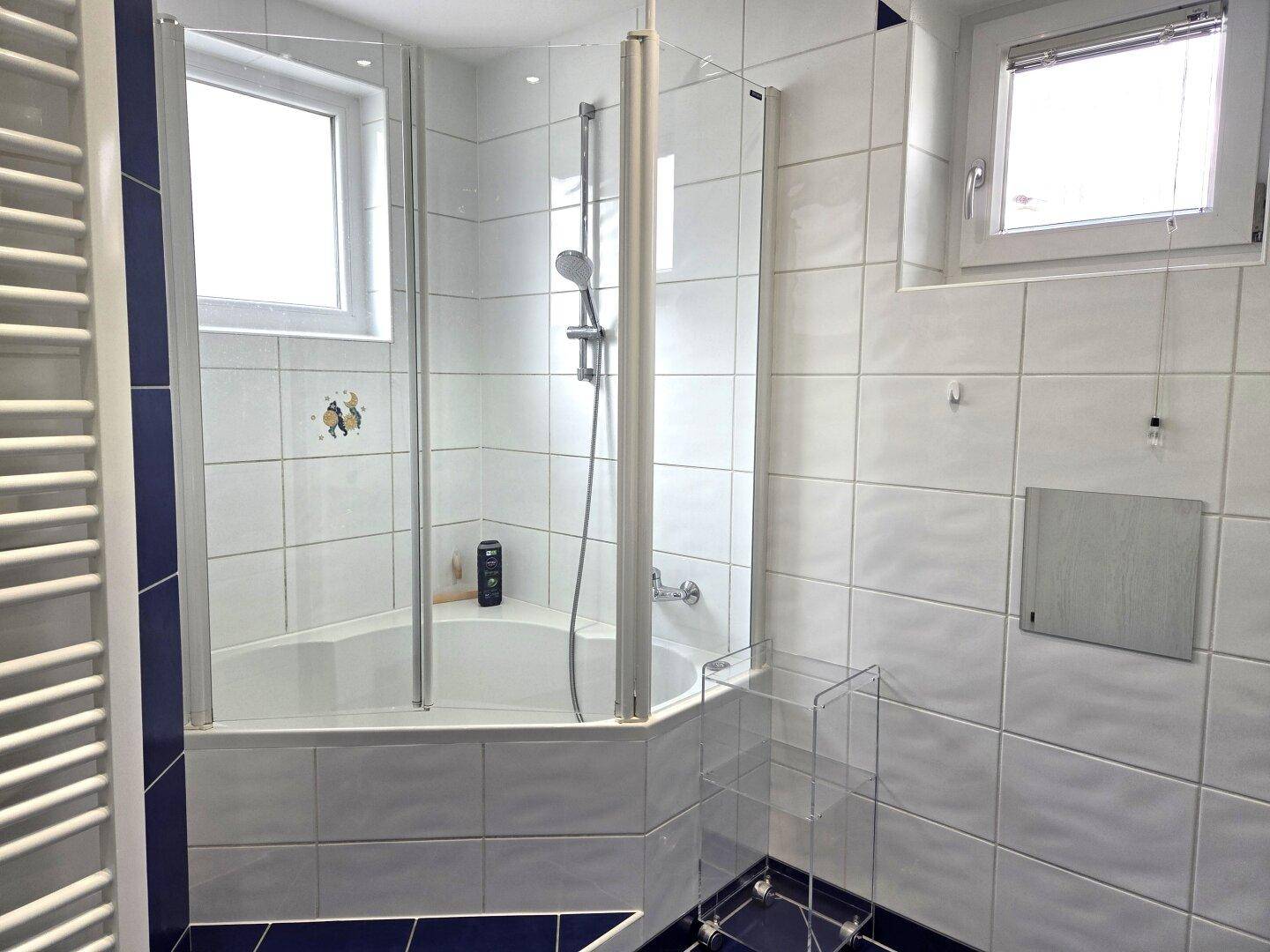 Badezimmer mit Eckbadewanne und integrierter Dusche, weißen Fliesen und einem Fenster.