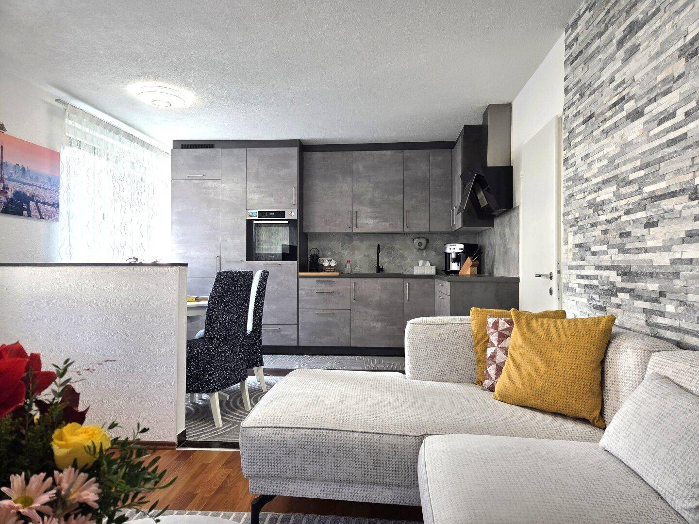 Moderne Küche mit grauen Fronten und integrierten Geräten, offen zum Wohnbereich mit Steinwand.