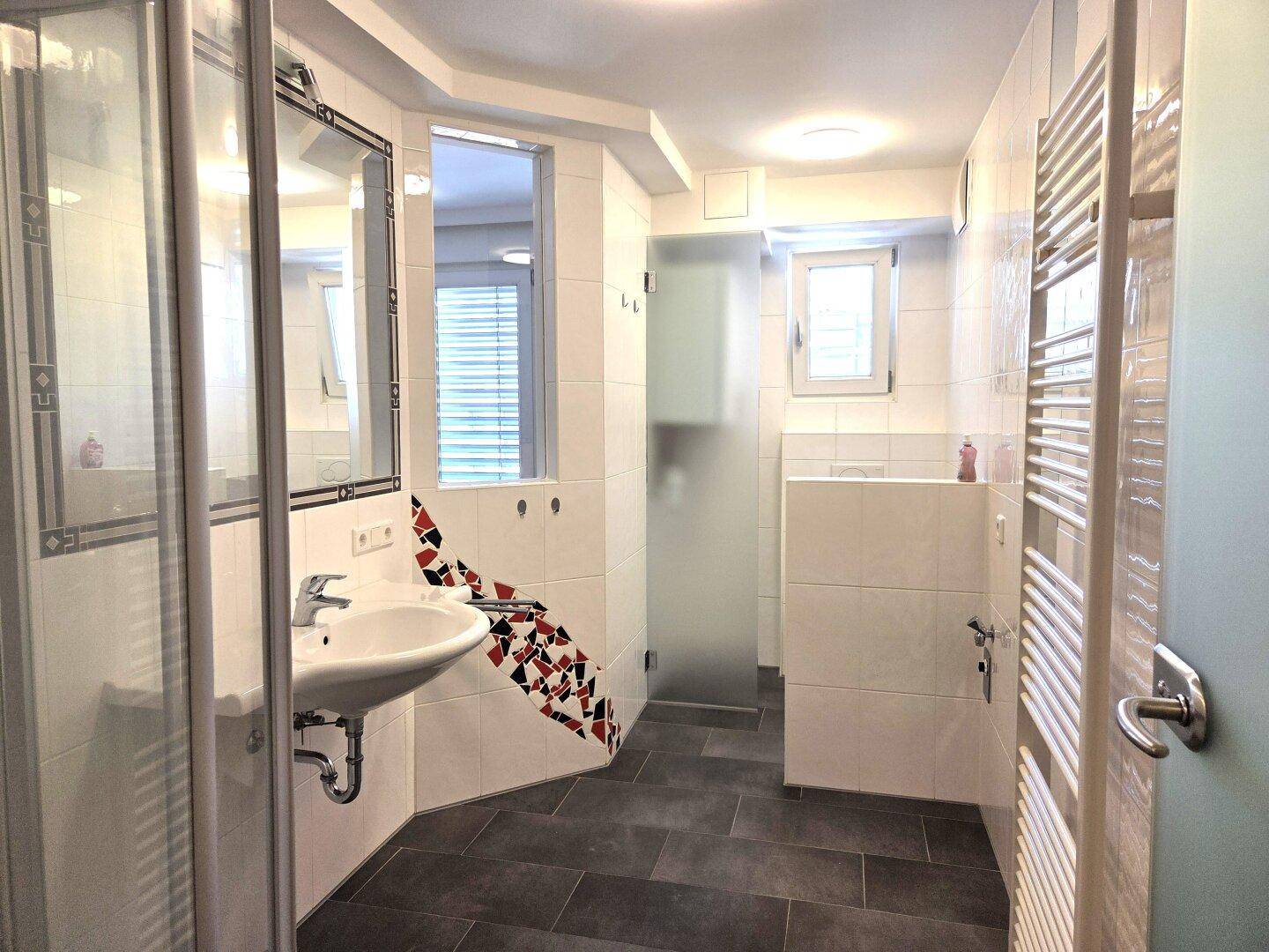 Modern gefliestes Badezimmer mit Dusche, Waschbecken, Spiegelschrank und Handtuchheizkörper sowie Fenster.