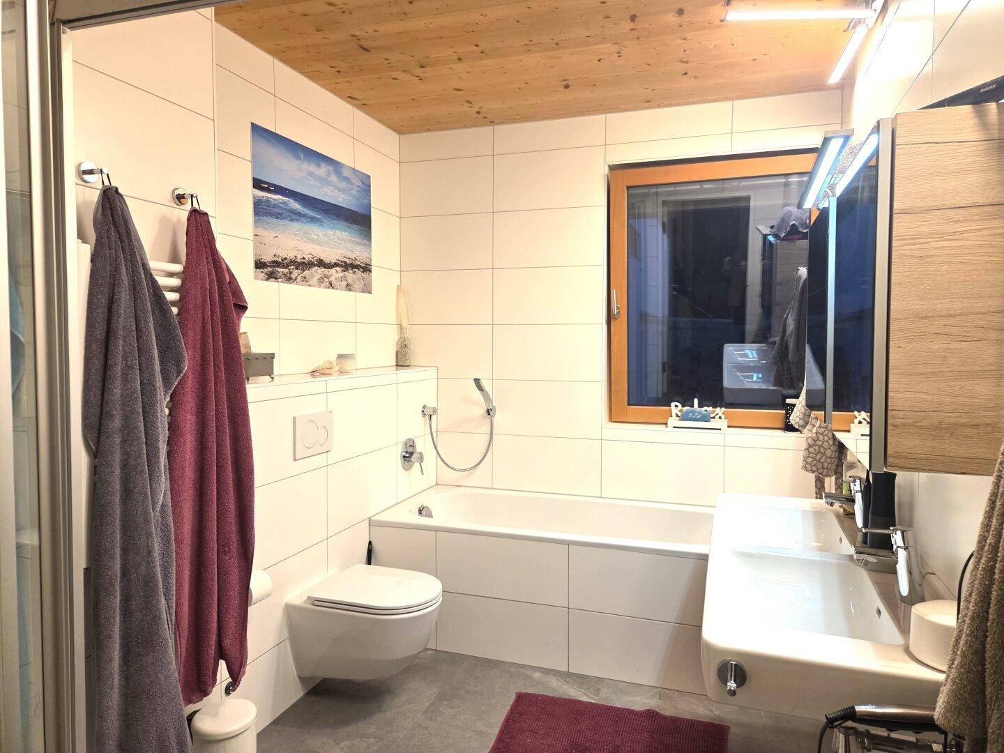 Modernes Badezimmer mit Badewanne, Wand-WC, großem Spiegel und einem Fenster für Tageslicht.