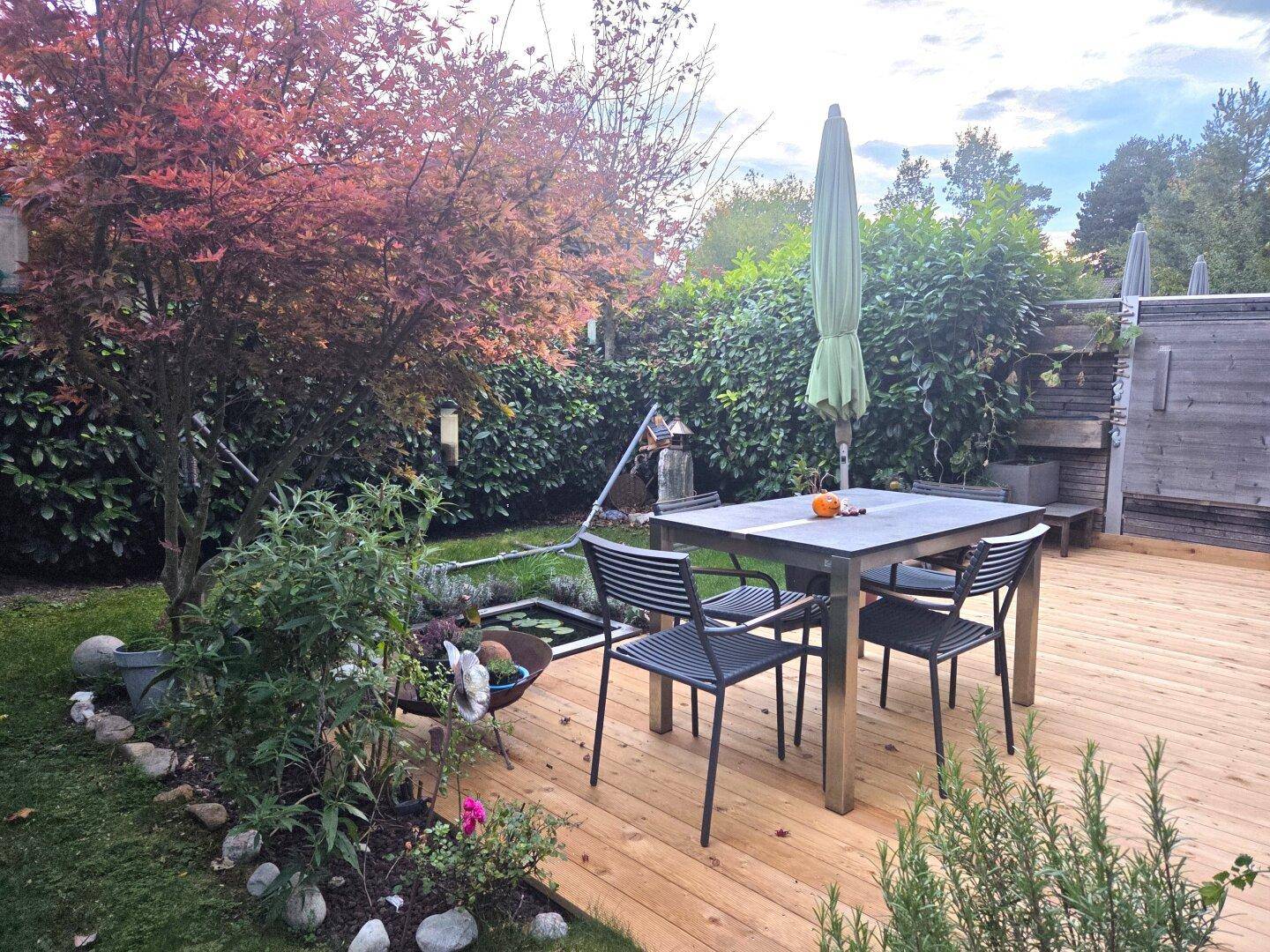 Sonnige Holzterrasse mit Esstisch, Stühlen und umgebender Bepflanzung, ideal für Mahlzeiten im Freien.