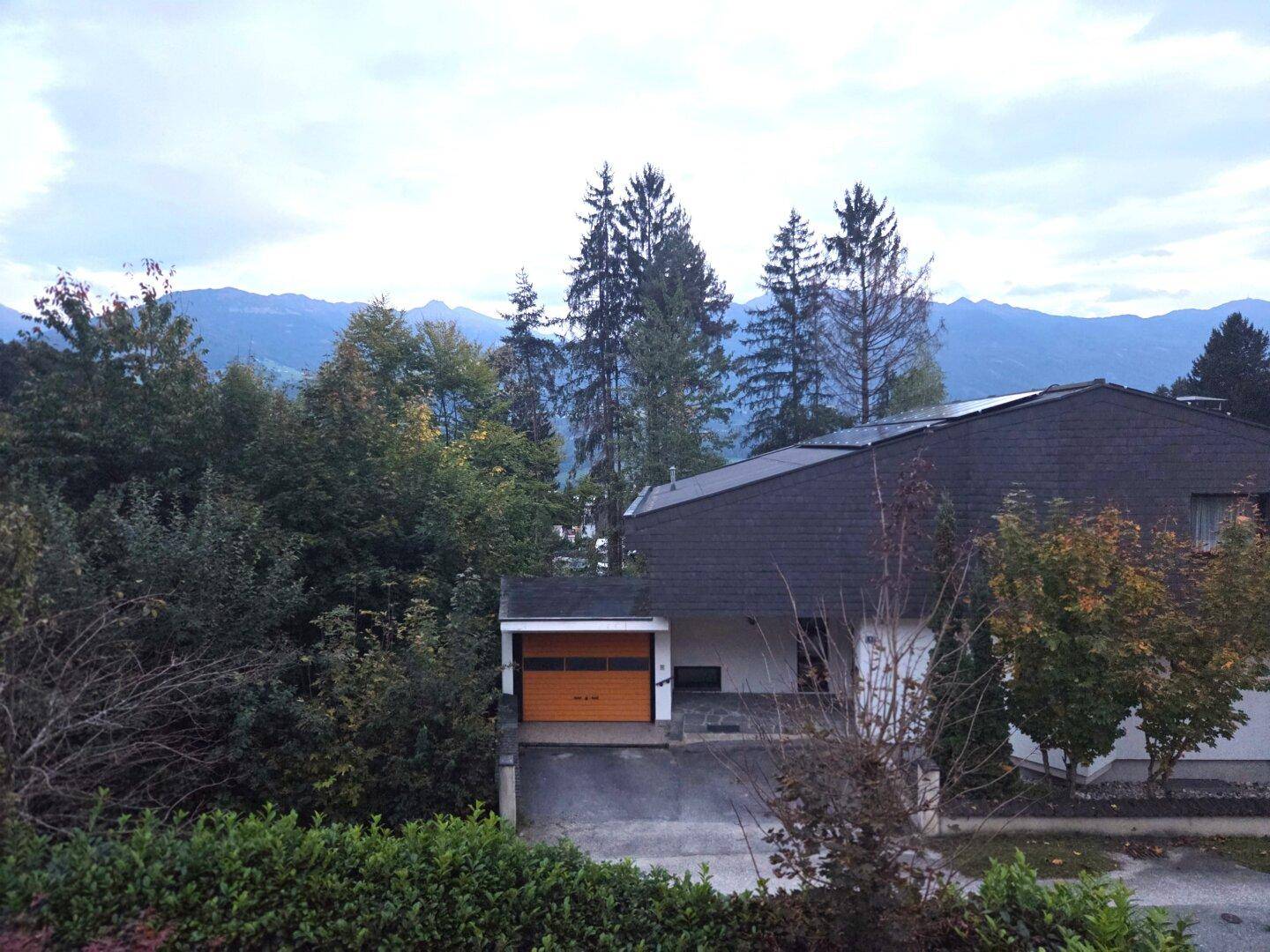 Hausfassade mit Garage und umgebender Natur, im Hintergrund sind Berge sichtbar.