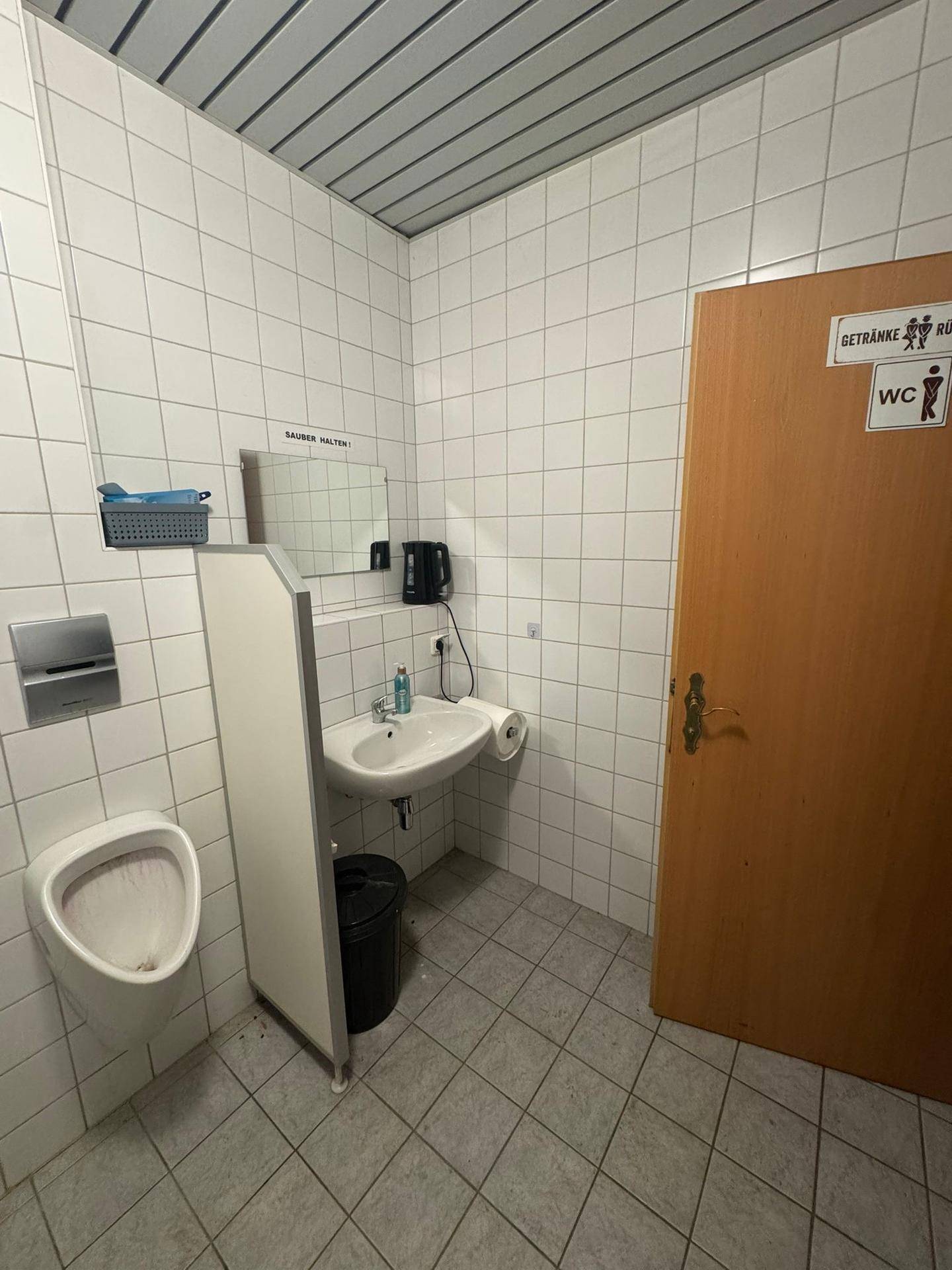 Gefliestes Badezimmer mit Urinal, Waschbecken und einer Holztür, in gutem und funktionalem Zustand.