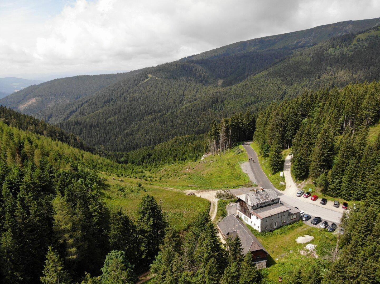 Luftaufnahme eines Gebäudekomplexes in einem bewaldeten Bergtal mit Straße und Parkplatz.