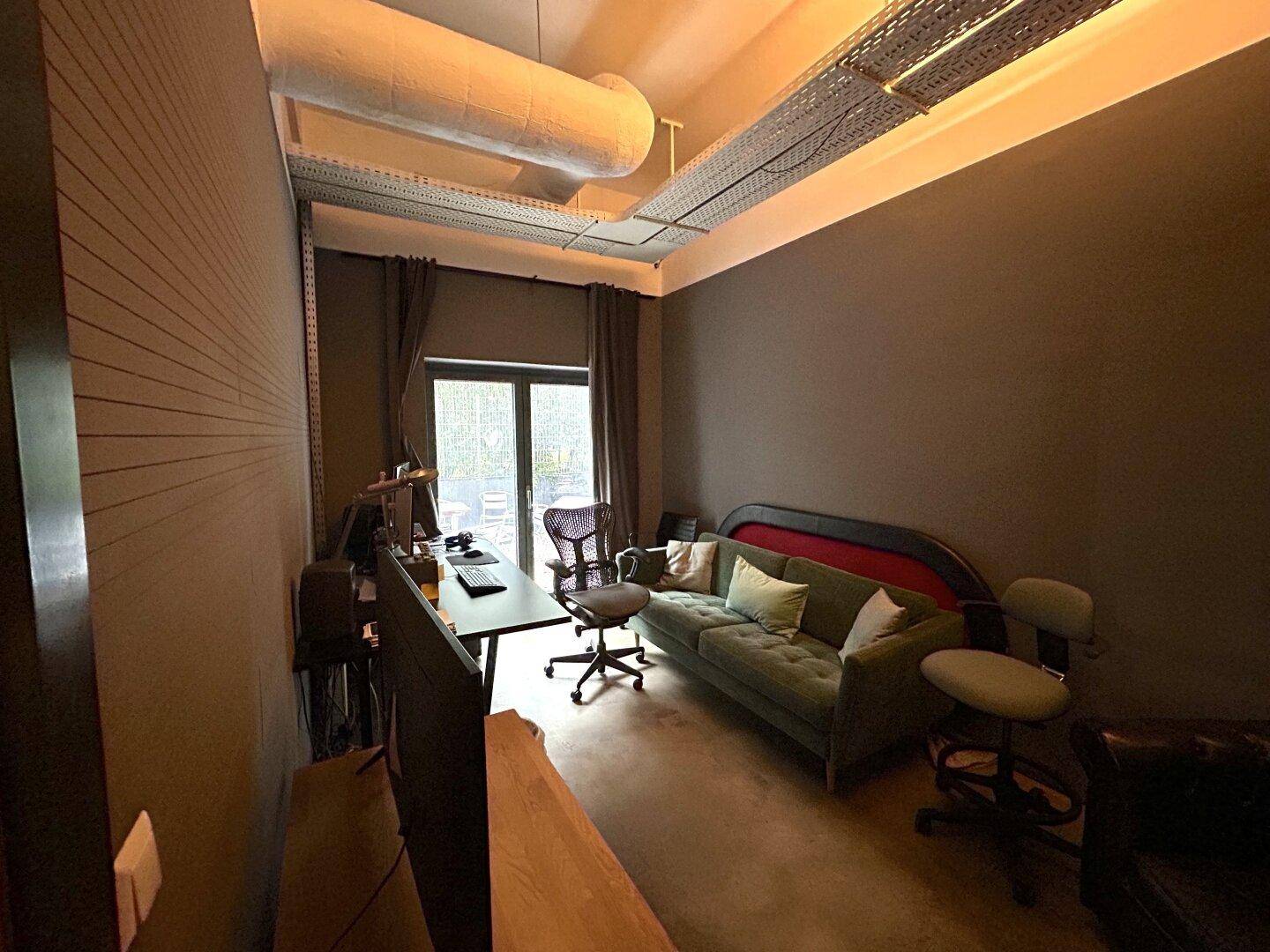 Dunkel gehaltener Büroraum mit Sofa, Arbeitsplatz, hohen Decken und industriellen Rohren.
