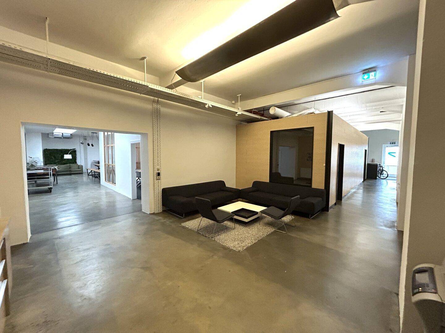 Geräumiger offener Bürobereich mit bequemen Sofas, Teppich und industriellem Charme durch sichtbare Leitungen.