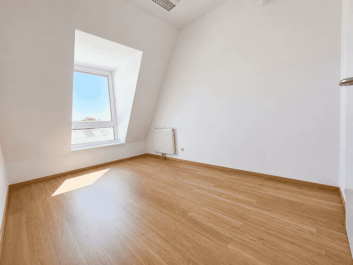 Leerer Raum mit weiß gestrichenen Wänden, Dachschräge und einem Fenster, ausgestattet mit Holzoptikboden und Heizkörper.