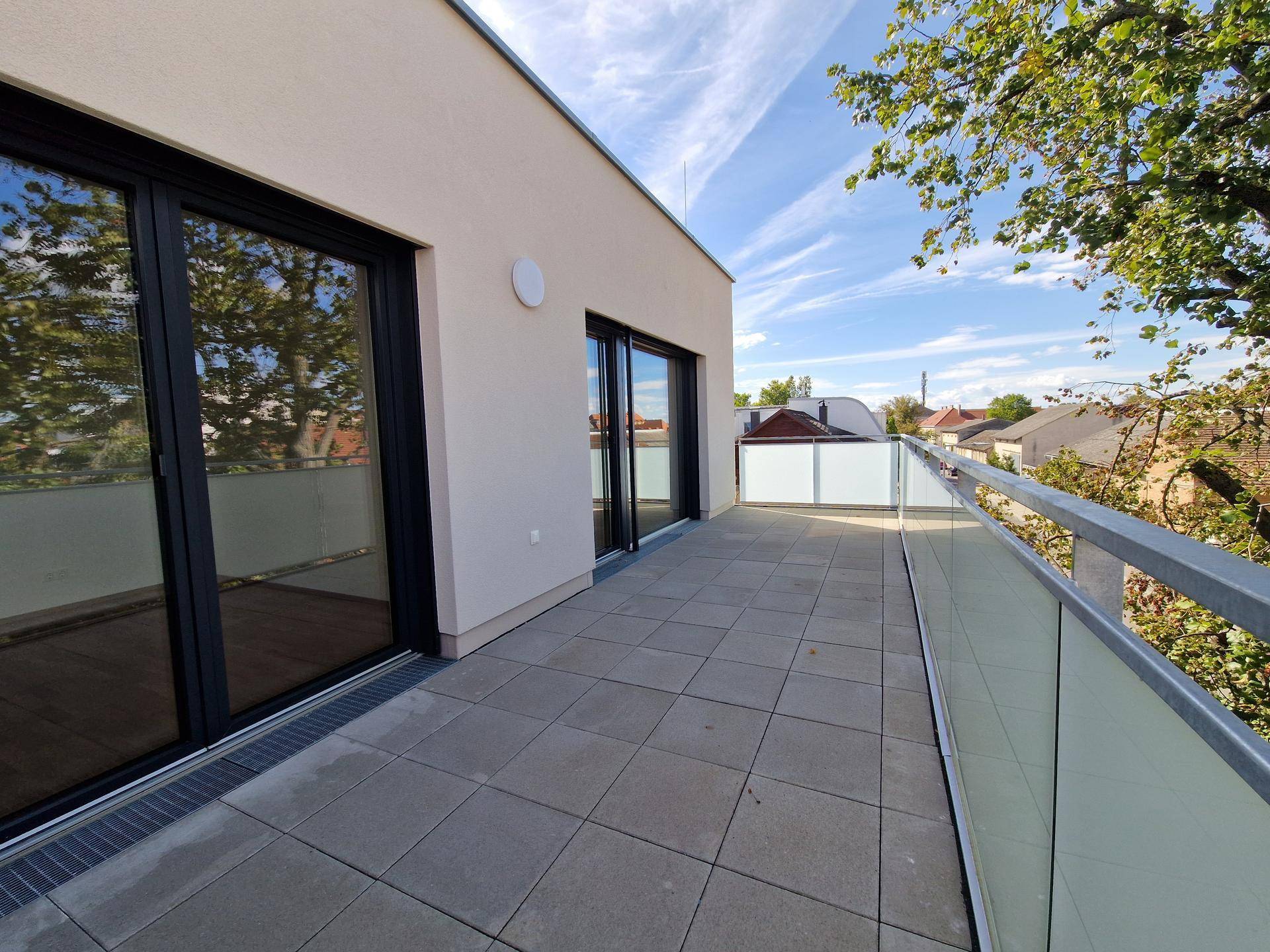 Geräumige Terrasse mit Fliesenboden, Glasgeländer und Zugang zu den Innenräumen, umgeben von Bäumen.