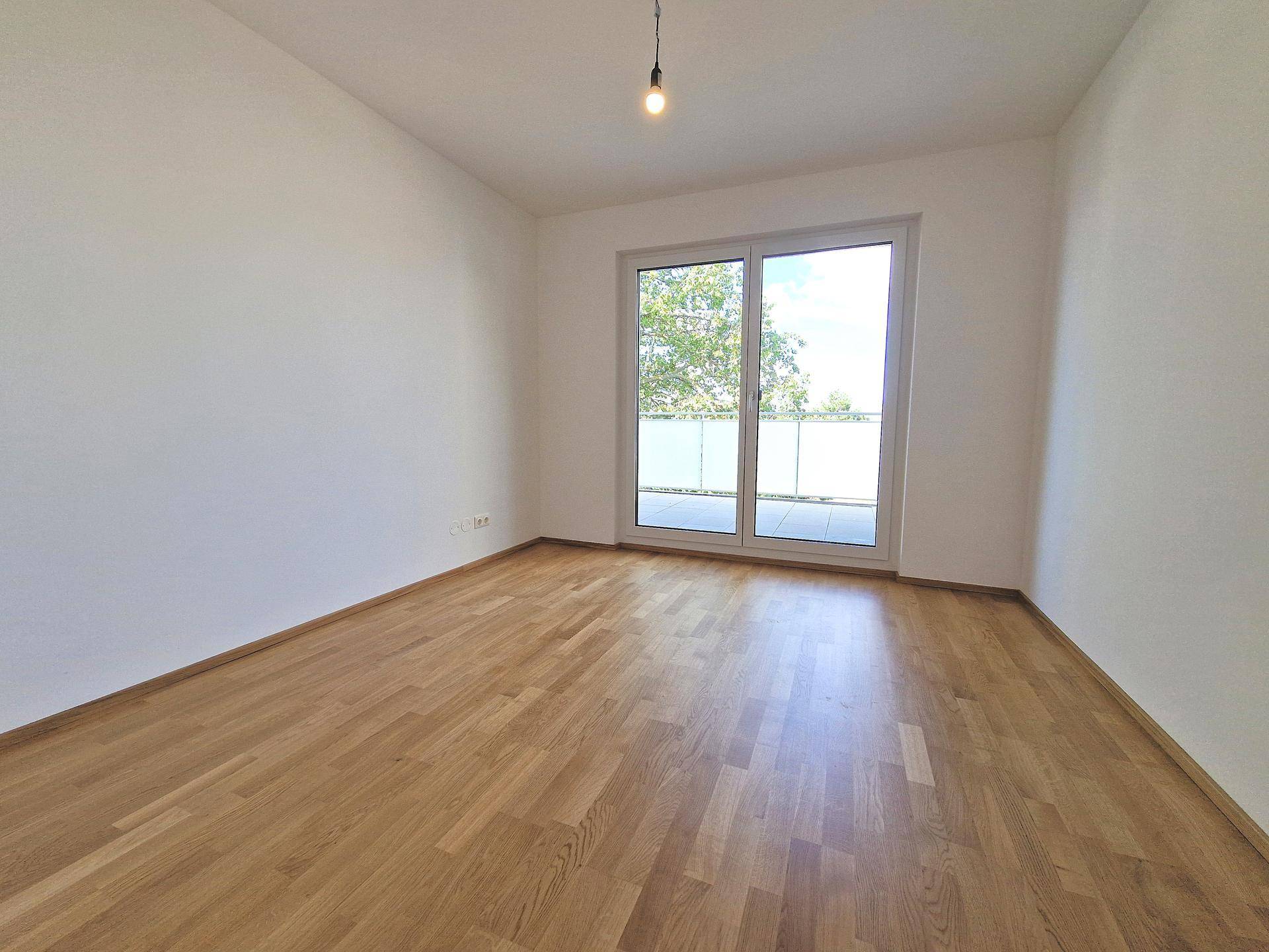 Leeres Schlafzimmer mit hellem Parkettboden, weißen Wänden und großem Fenster mit Zugang zur Terrasse.