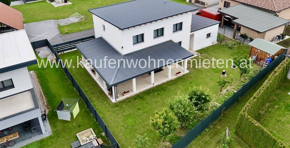Modernes Einfamilienhaus mit weißer Fassade, dunklem Dach und gepflegtem Garten mit überdachter Terrasse.