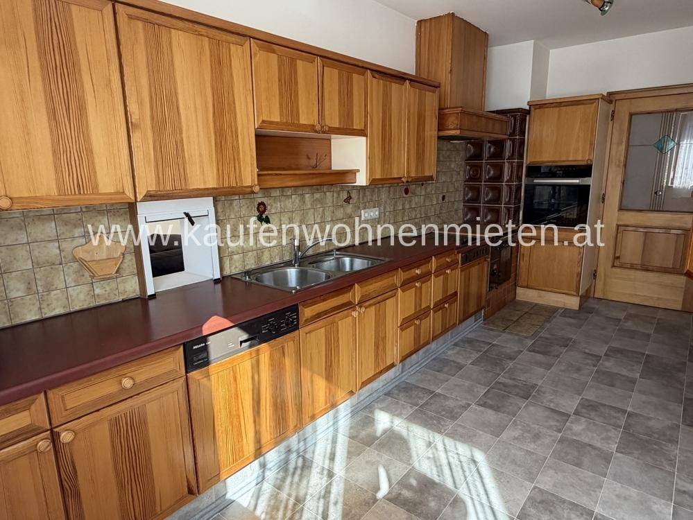 Traditionelle Holzküche mit Fliesenboden, Einbaugeräten und einem Kachelofen im Hintergrund.