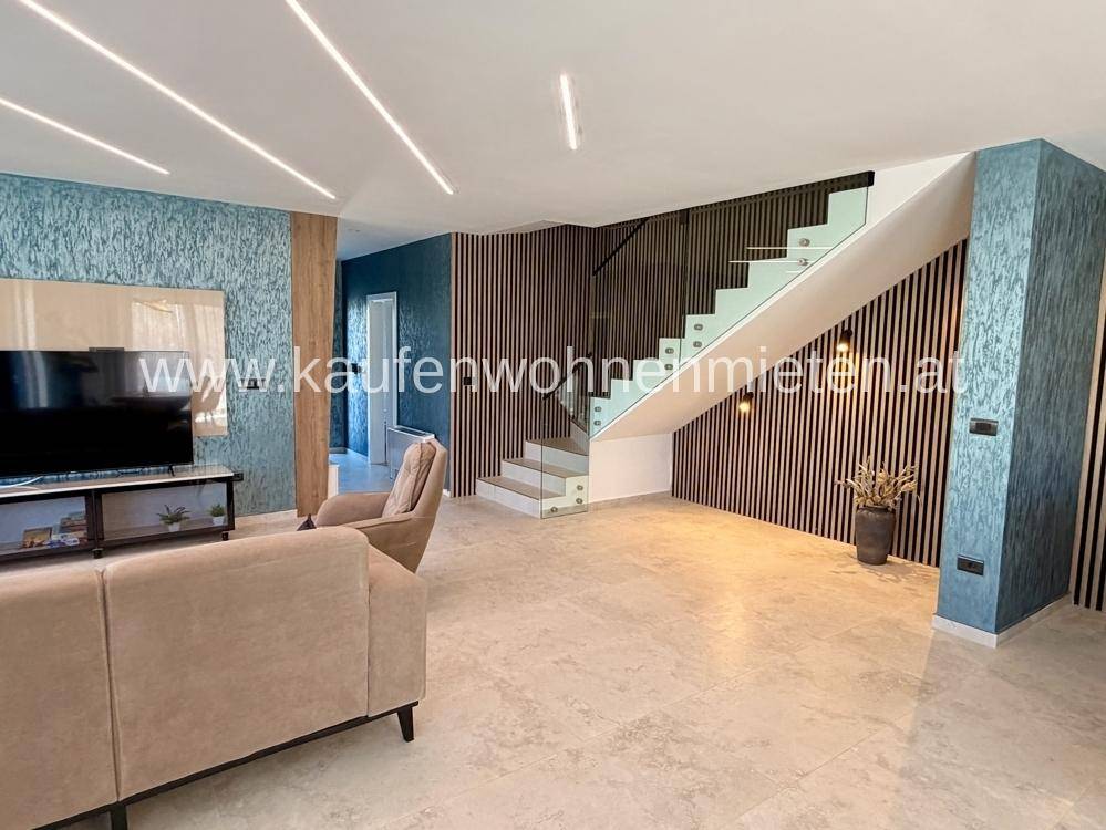 Geräumiges Wohnzimmer mit heller Fliesenboden, beigem Sofa und moderner Treppe mit Glasgeländer.