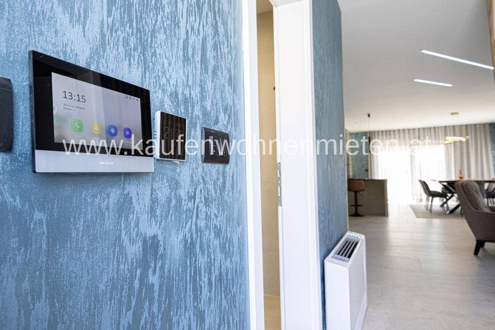 Smart-Home-Bedienfeld an einer strukturierten blauen Wand mit Blick in den Wohnbereich.
