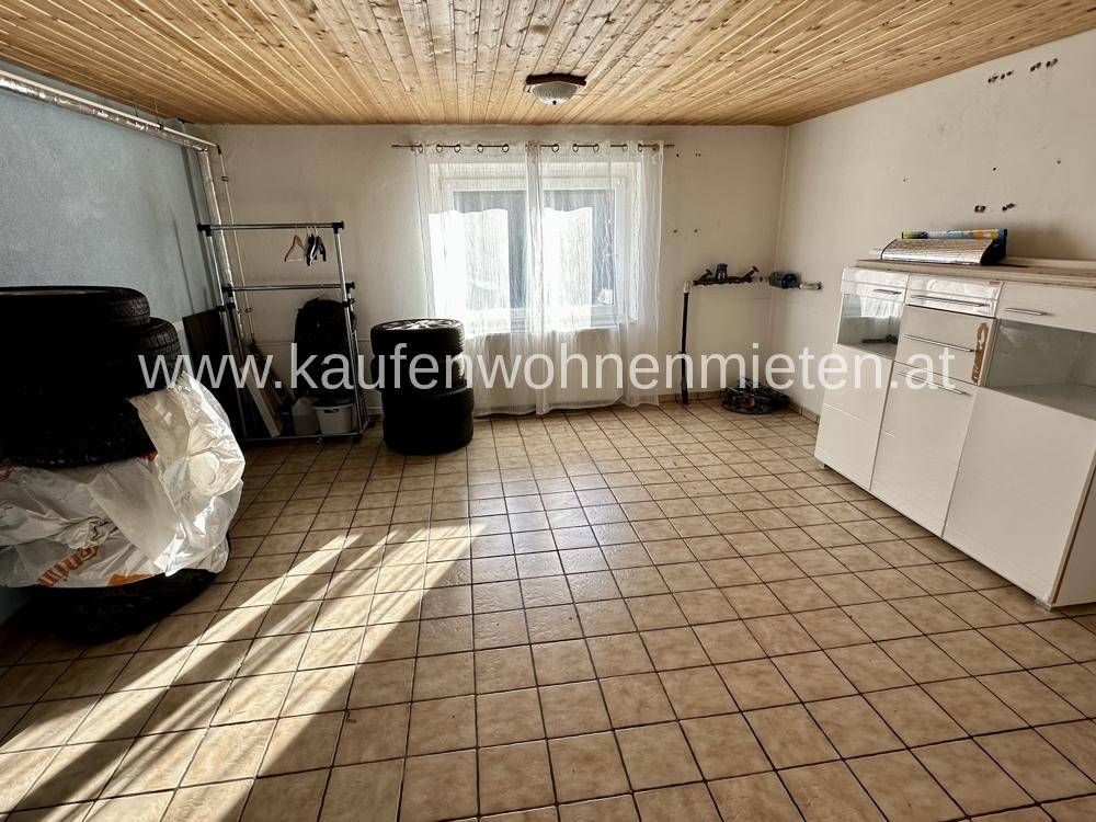 Geräumiger Raum mit Fliesenboden, Holzdecke und Fenster, derzeit als Lager genutzt.