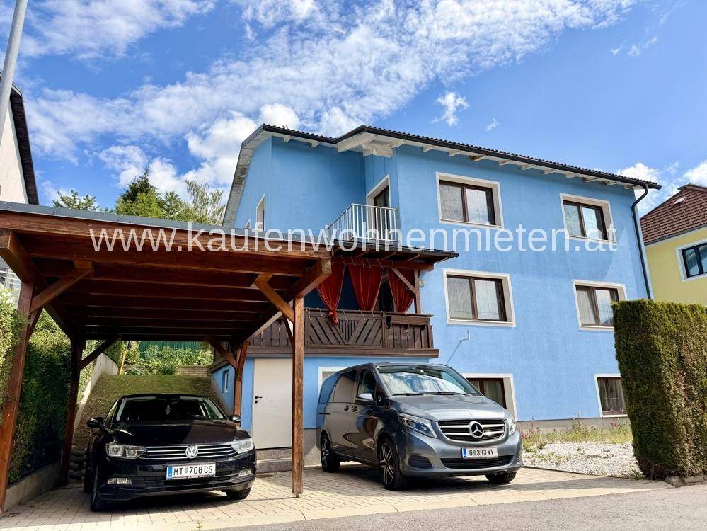 Blaues Wohnhaus mit überdachtem Carport, Stellplätzen und kleinem Balkon.