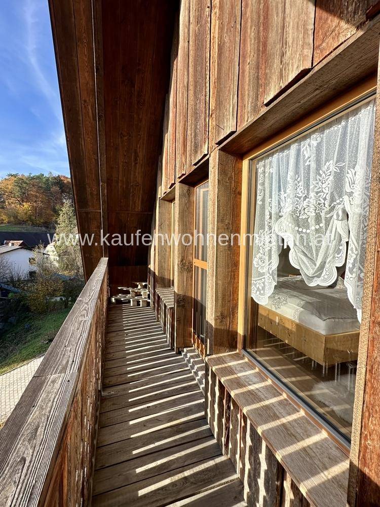 Langer Holzbalkon mit Blick ins Grüne und traditionellen Spitzenvorhängen am Fenster.