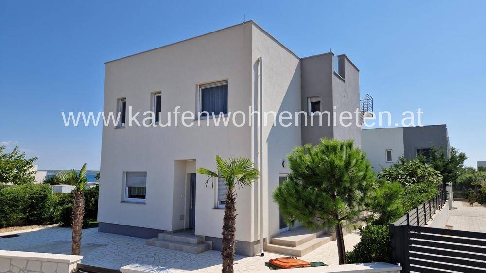 Moderne weiße Hausfassade mit klaren Linien und kleinen Palmen im Vorgartenbereich.