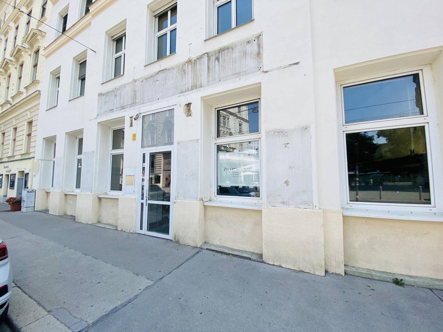 Die Erdgeschossfassade des Gebäudes mit Eingangstür und großen Fenstern, die leichte Abnutzungserscheinungen aufweist.