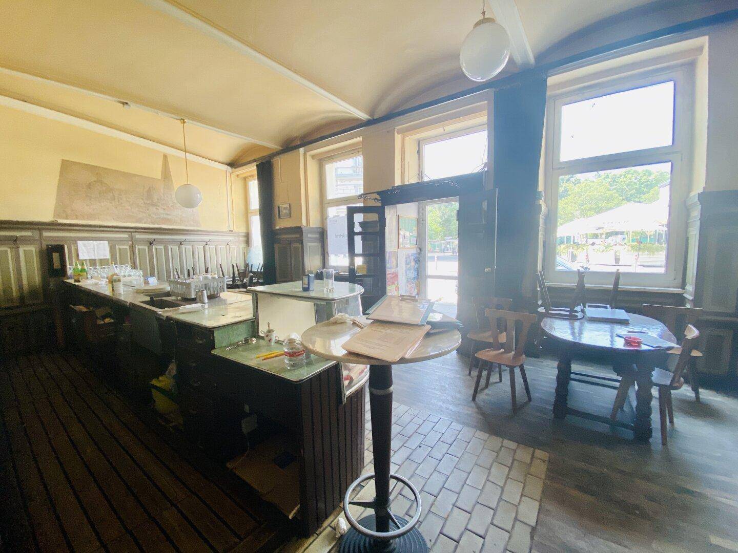 Der Barbereich eines Restaurants mit Theke, Hockern und Tischen, ergänzt durch große Fenster und unterschiedliche Bodenbeläge.