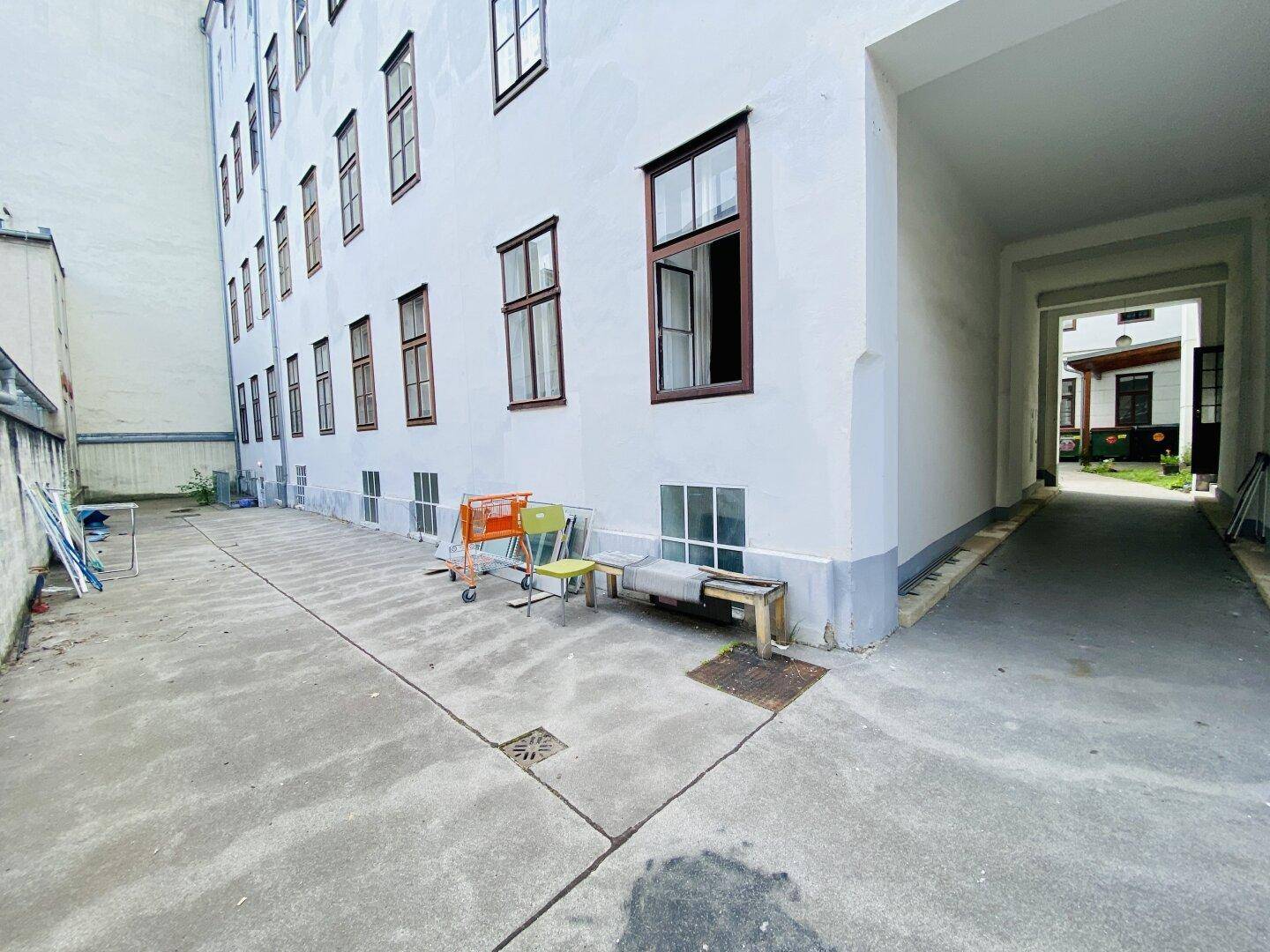 Gepflasterter Innenhof neben einem weißen Gebäude mit einem Bogendurchgang zu einem weiteren Bereich.